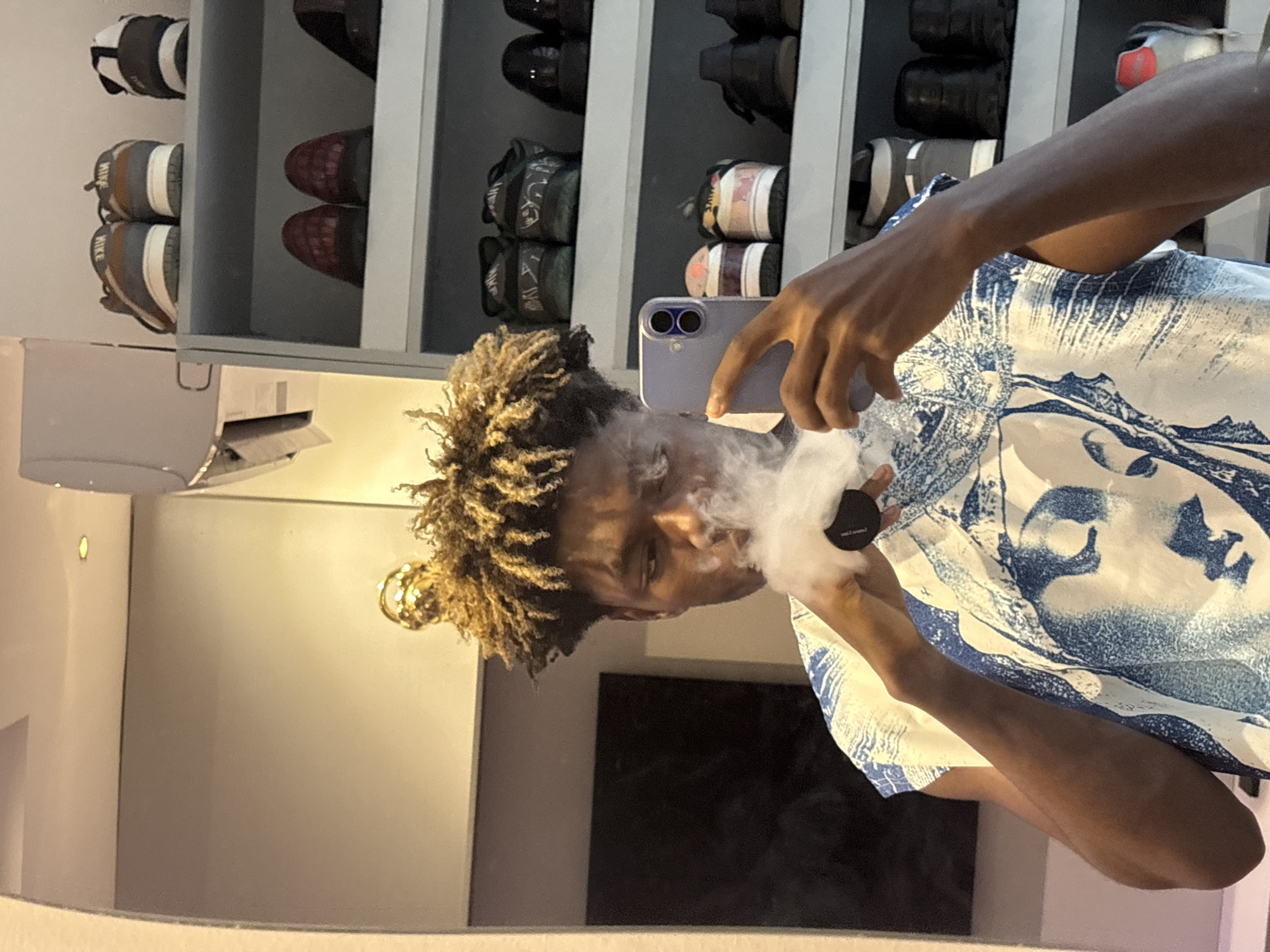 A medium shot of a person smoking from a device in front of a mirrored wall. The person has short dreadlocks and is wearing a blue and white shirt. The smoke obscures the person's face. The person is holding a light purple phone in front of their face. Behind the person is a shoe rack containing various pairs of shoes. An air conditioning unit is attached to the wall. A black rectangular object is also visible. No information to identify the location in Igboefon, Nigeria.