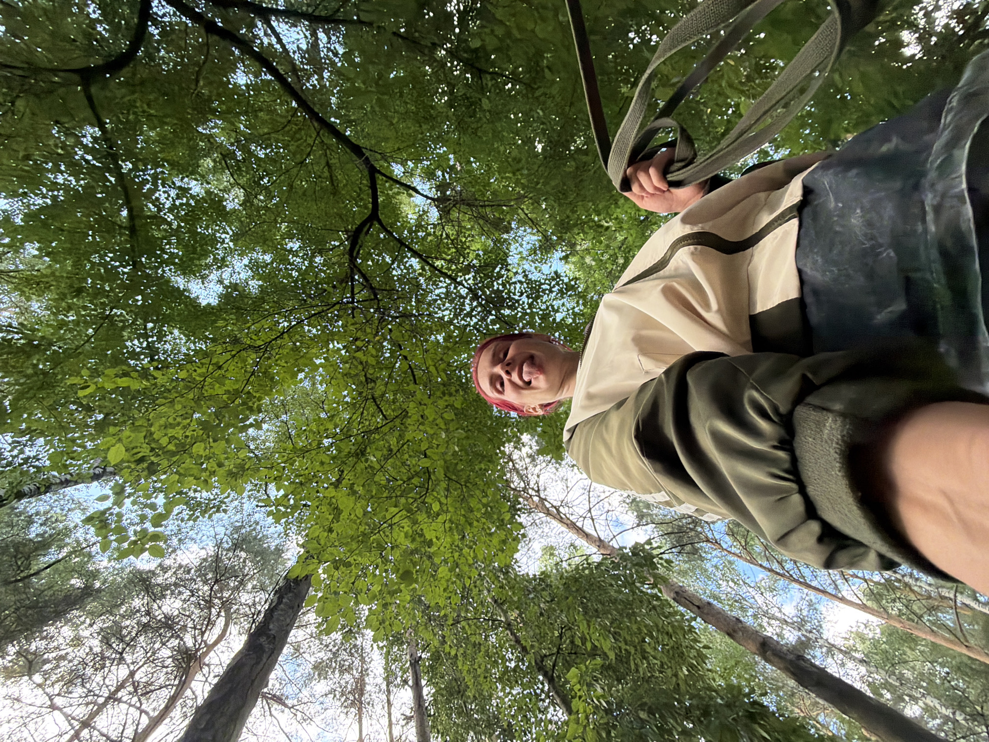 An individual with brightly colored hair sticks out their tongue towards the camera, while holding a coiled green strap. The person is wearing a light-colored jacket with green accents and a dark-colored skirt. They are positioned outdoors within a forest, with a canopy of green leaves and tall trees overhead. The perspective is from below, looking up at the person and the sky. The scene is devoid of any urban elements, suggesting it is not in the city of Chaikovsky, Russia.