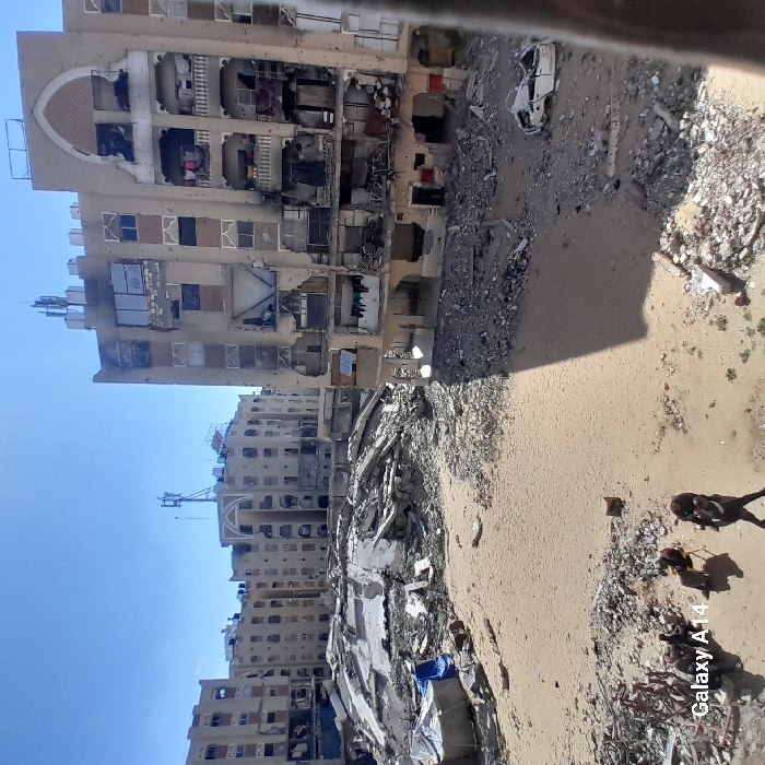 The image depicts a severely damaged urban environment in Khan Yunis, Palestinian Territory, under a clear blue sky. The foreground shows a sandy expanse mixed with extensive rubble from collapsed structures. In the lower-left, two individuals are present on the sandy ground; one person in dark clothing walks to the right, while another is seated on a yellow chair next to a dark object. A blue tarp structure is visible to the left. To the right, a heavily damaged white vehicle is partially buried in debris. The middle ground is dominated by large piles of concrete slabs, twisted rebar, and other construction materials, indicative of entirely collapsed buildings. The background features multiple multi-story residential buildings, all exhibiting significant structural compromise. Damage includes large holes through facades, missing wall sections, exposed interiors, shattered windows, and charring. One prominent building on the right displays an arched decorative element on its upper facade. Rooftops feature visible communication antennas. Strong sunlight casts distinct shadows across the scene.
