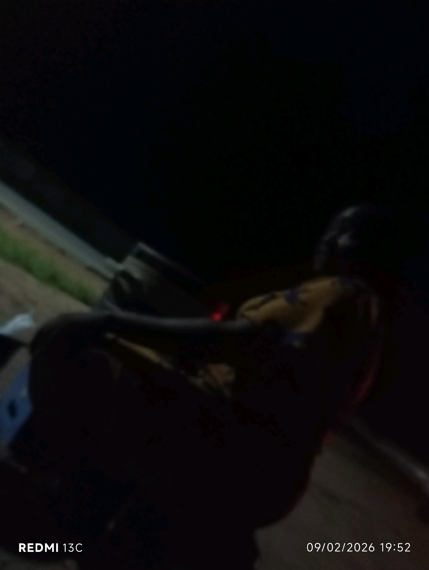 A dimly lit, slightly blurry image shows a person reclining in a dark environment, possibly outdoors at night. The person is wearing a yellow and purple patterned garment. To the left of the person, a light-colored object, possibly a bag, is partially visible. The background is dark, with a hint of greenery on the left. The lower left corner of the image displays "REDMI 13C" and the lower right corner shows a date and time: "09/02/2026 19:52". The context provided suggests this scene is located in the city of Shendam, Nigeria.