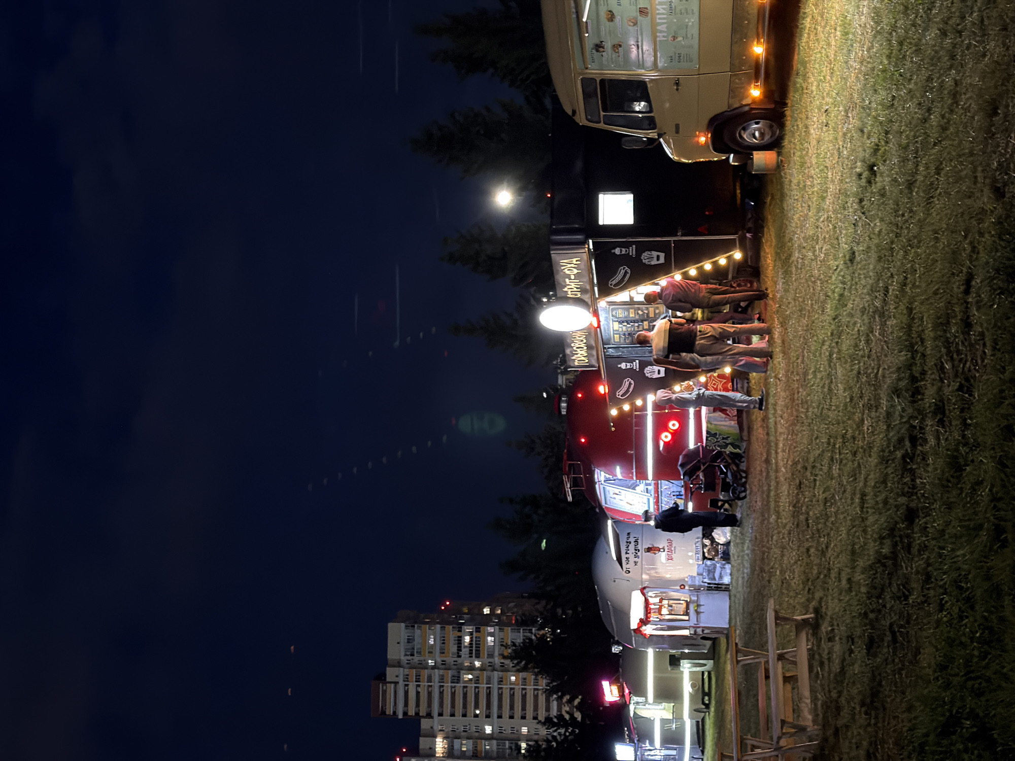 The image depicts a nighttime scene at what appears to be a food truck festival or outdoor market. Multiple food trucks are lined up, illuminated by string lights and the interior lights of the trucks. The ground is covered in grass, suggesting an outdoor park or open field setting. In the background, a multi-story apartment building with illuminated windows is visible, indicating an urban or suburban environment.
Several people are present, interacting with the food trucks. Some are standing in line, while others appear to be ordering or receiving food. The mood seems casual and social, typical of an evening gathering. The sky is dark, with a bright moon visible, suggesting a clear night.
One of the food trucks has text on its side that reads "СПИРТ-ФУД," which translates to "SPIRIT-FOOD." Another truck has text that appears to say "ОТ НАС РОЖДЕН" and "НЕ ПРОБЛЕМА," roughly translating to "BORN FROM US" and "NOT A PROBLEM." The truck at the top of the image has a menu displayed with items like "НАПИТКИ" (DRINKS) and "КЛАССИК" (CLASSIC).
The scene is lit by artificial lights, creating a warm and inviting atmosphere despite the darkness of the night. The combination of food trucks, people, and the urban background creates a lively and engaging atmosphere.