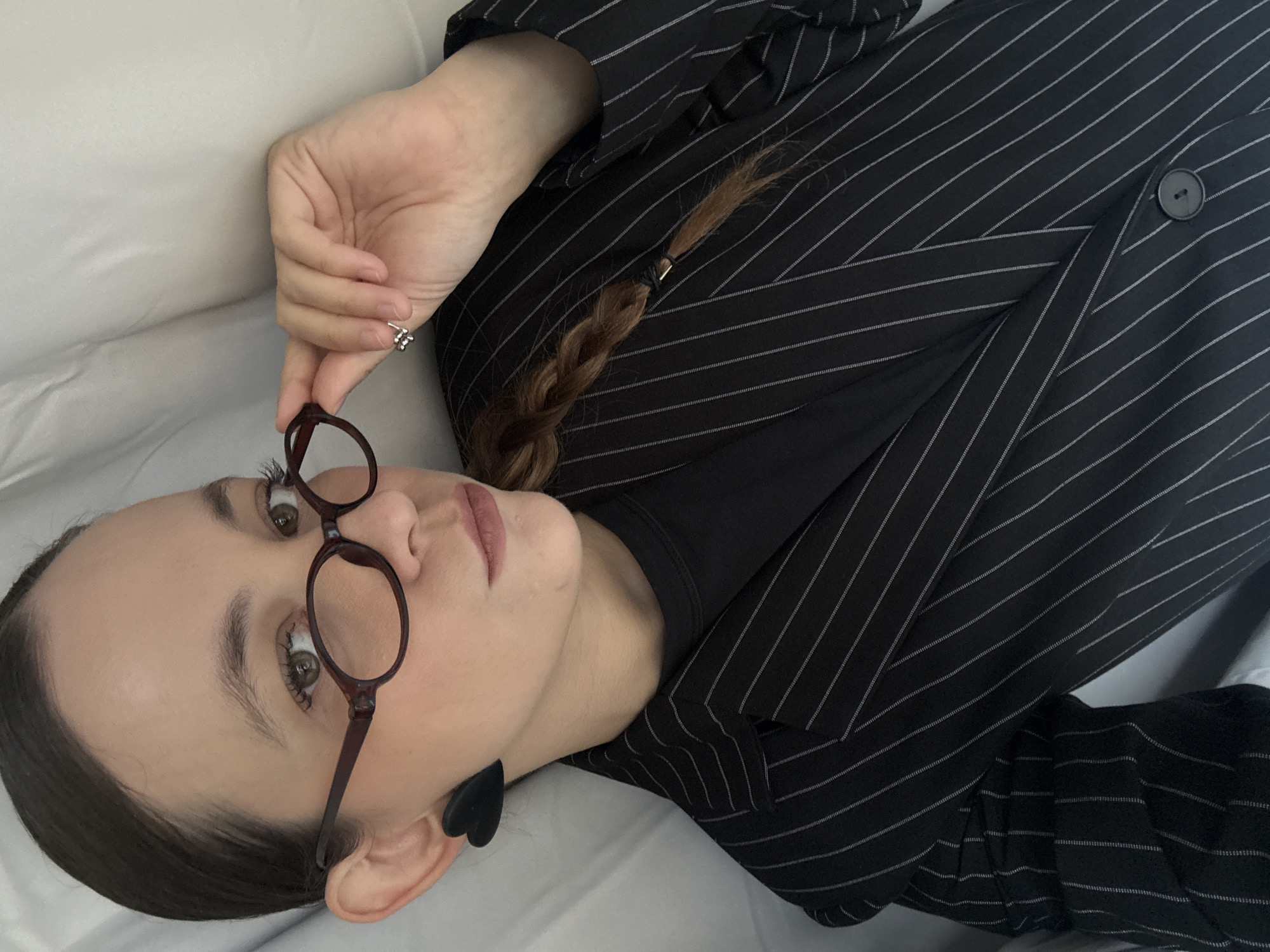 A young woman with light skin and brown hair styled in a braid is lying down on a white surface. She is wearing a black pinstripe blazer and a black top. The woman is holding a pair of round, dark-rimmed glasses up to her face, looking towards the camera. She has on small, silver stud earrings, one of which is visible on her right hand. Black heart-shaped earrings are also visible. The lighting suggests it is daytime, and the indoor setting is implied by the white surface, which could be a bed or couch. The woman's expression is neutral. There is no visible text in the image.