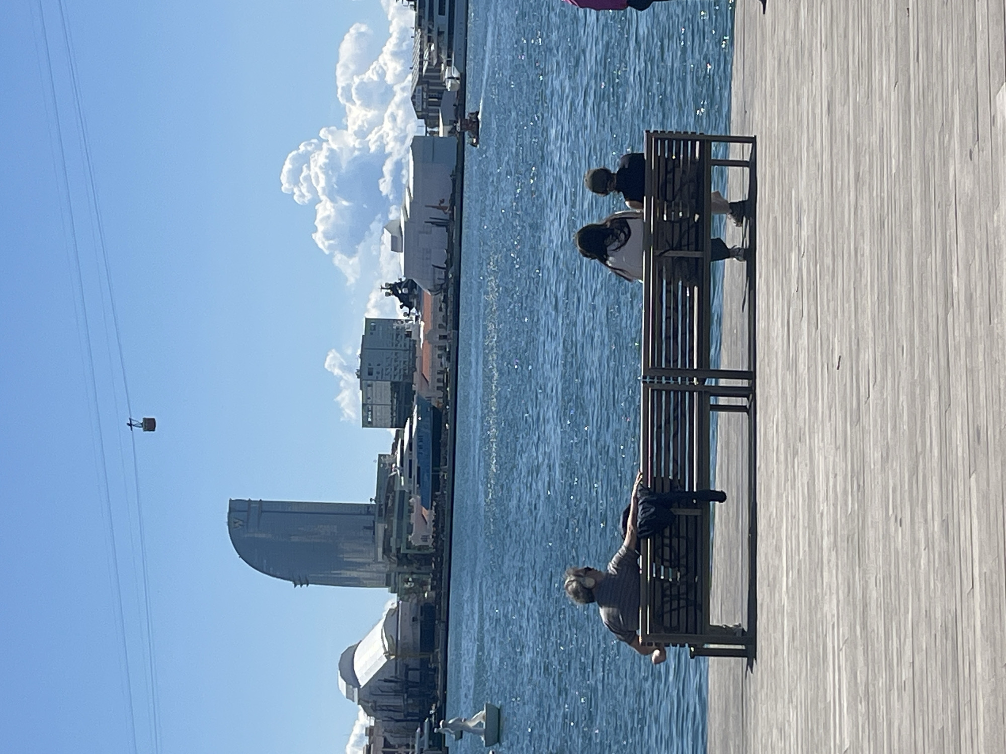 The image depicts a waterfront scene in Barcelona, Spain. In the foreground, people are sitting on benches along a wooden boardwalk overlooking the harbor. To the left, the Hotel Arts Barcelona and other buildings are visible, with a cable car ascending towards Montjuïc. The harbor is filled with water reflecting sunlight, and white clouds dot the sky.