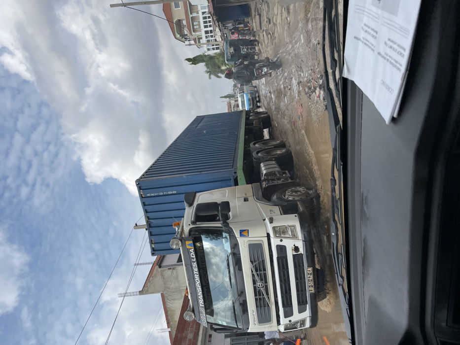 The image captures a street scene in Luanda, Angola, from the perspective of a vehicle's dashboard. The main subject is a large white Volvo truck with a blue shipping container loaded on its trailer, positioned on a muddy, unpaved road. The truck's cab has "VOLVO" prominently displayed on its grille, and its license plate "LD-51-42-EA" is visible. Text on the sun visor reads "RUA TRANSPORTES LDA". Part of the container's identification "ARTU 700" is also visible.
The road is extensively covered in large puddles and mud, suggesting recent rain. In the background, other vehicles, including a motorcycle with a rider, navigate the challenging terrain. Several people are visible in the distance and near the truck's front wheel. Buildings, some appearing residential or commercial, line the sides of the street, with a cloudy sky overhead. Overhead wires are also noticeable. The time of day is daytime, with bright but overcast conditions.
Inside the viewing vehicle, a white document with printed text is taped to the dashboard or windshield. Visible words on the document include "CONTRACTO" and a number "500.000". The scene depicts a bustling, everyday urban environment characterized by difficult road conditions.