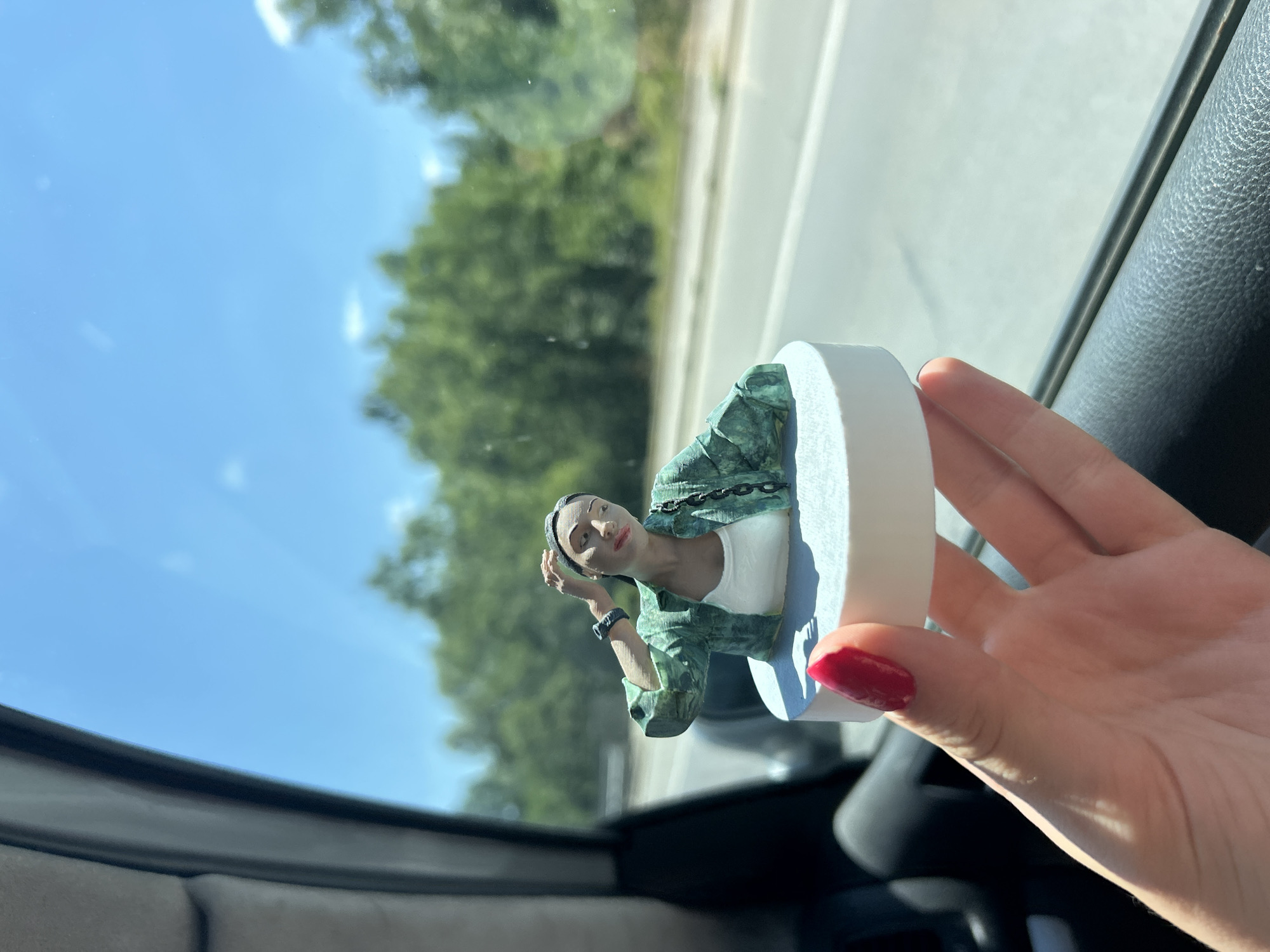 A close-up, eye-level shot shows a hand with bright red nail polish holding a small, 3D-printed figurine of a woman. The figurine, which appears to be a miniature replica of a person, is wearing a green patterned shirt over a white tank top and a dark watch on its left wrist. It is positioned on a white circular base. The background, seen through a car window, features a bright blue sky with scattered clouds and lush green trees lining a paved road, suggesting an outdoor setting likely outside of Sofia, Bulgaria, possibly on a highway or main road during daylight hours. The interior of the car is visible on the left side of the frame, with dark gray textured material forming the car door. The overall lighting indicates a sunny day.