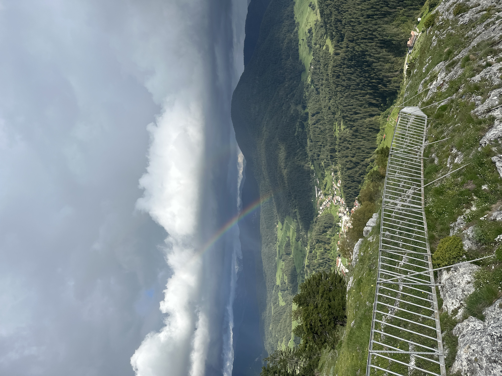 The image presents a landscape view from a high vantage point, featuring a metal grate walkway along a rocky cliff edge. Below the cliff, a lush green valley is visible, dotted with a small village or town. A mountain range forms the backdrop. In the sky, there is a dramatic interplay of dark storm clouds and brighter patches, with a segment of a rainbow visible across the valley. The scene appears to be in a mountainous region, but there is no indication of it being Iagodina, Bulgaria. No people, actions, or interactions are depicted. The primary subjects are the landscape, the rainbow, and the metal structure.