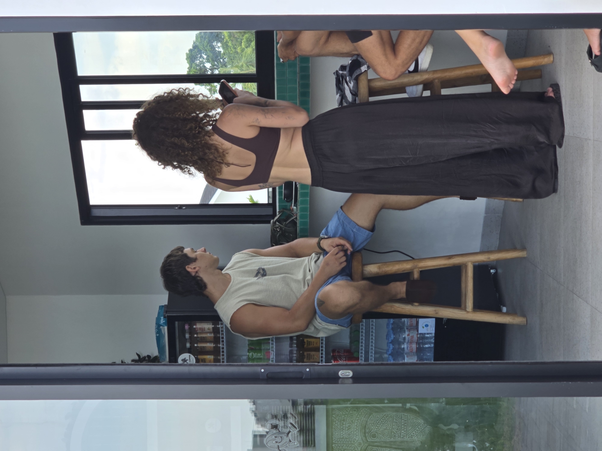 The scene shows a partially obscured indoor space in what appears to be North Kuta, Indonesia. A man with dark hair and a light tan tank top sits on a wooden stool in front of a glass-fronted refrigerator stocked with beverages. A woman with curly brown hair, wearing a dark sports bra and a long black skirt, stands nearby, holding a smartphone. Another person's legs are visible, seated on a wooden stool near a tile-covered wall. The view is partially framed by a glass sliding door, reflecting the exterior environment.