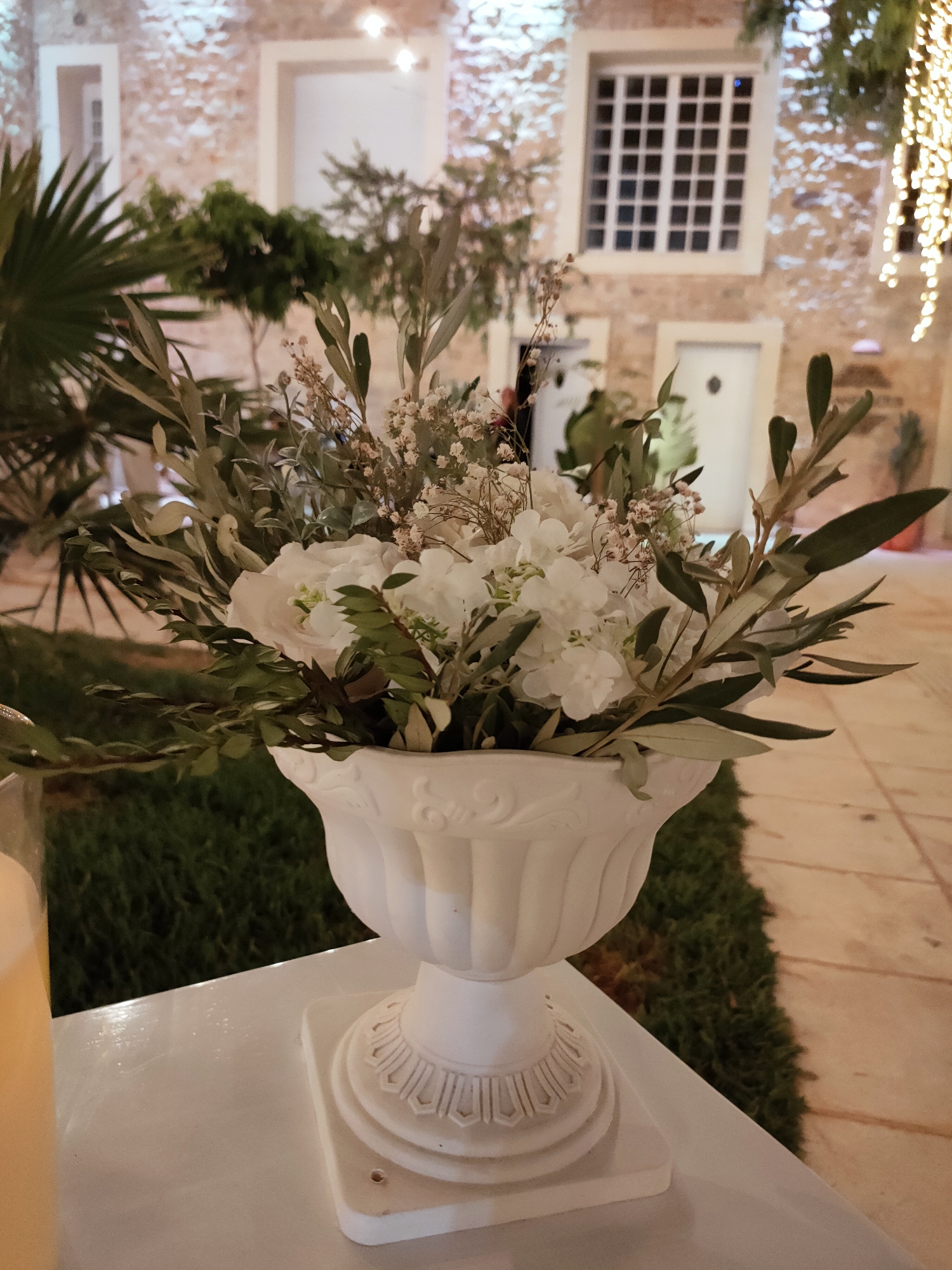 The image depicts a close-up of a white ornate vase filled with a floral arrangement. The arrangement consists primarily of white flowers, possibly roses and hydrangeas, interspersed with olive branches and smaller, delicate white flowers. The vase itself is intricately detailed with relief patterns. The vase is placed on a white surface, possibly a table, in what appears to be an outdoor setting.
In the background, a stone building with a rustic facade is visible. The building has multiple windows, including a large, grid-like window, and two arched doorways. Greenery, including palm fronds and other plants, frames the scene. String lights, with a warm glow, are also visible hanging in the upper right portion of the image, suggesting an evening or nighttime setting. The ground surface appears to be a combination of grass and cobblestone or paved flooring. The overall ambiance is one of an elegant outdoor venue, potentially for an event. The location is identified as Cebalat, Tunisia. There are no people visible in the image.