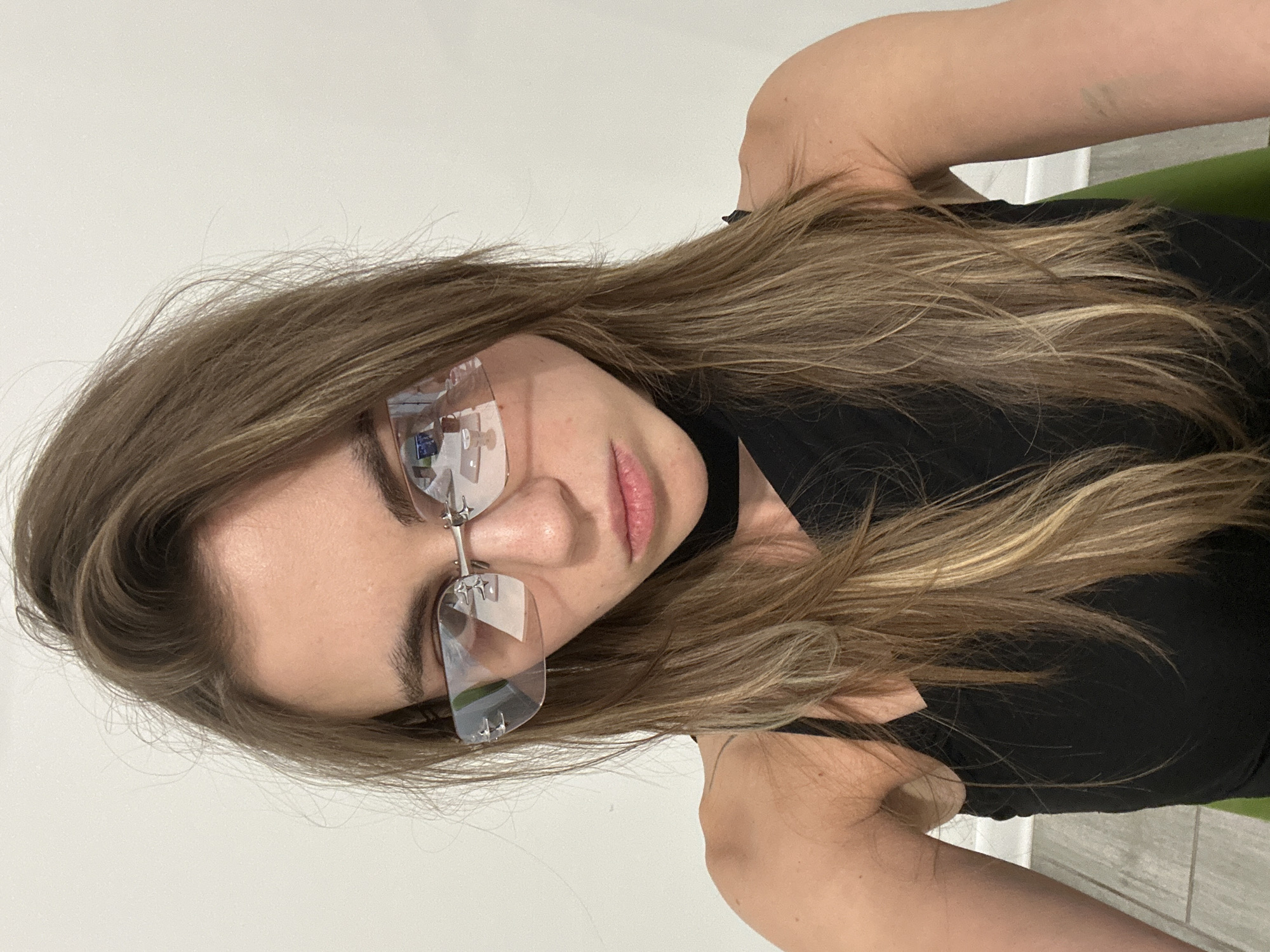 A young woman with long, light brown hair and fair skin, wearing rimless sunglasses and a black top, looks directly at the camera. Her lips are slightly pursed. The background is a plain white wall, with the suggestion of a tiled floor and a green object out of focus at the bottom. There are no discernible features that place the image in Moscow, Russia.