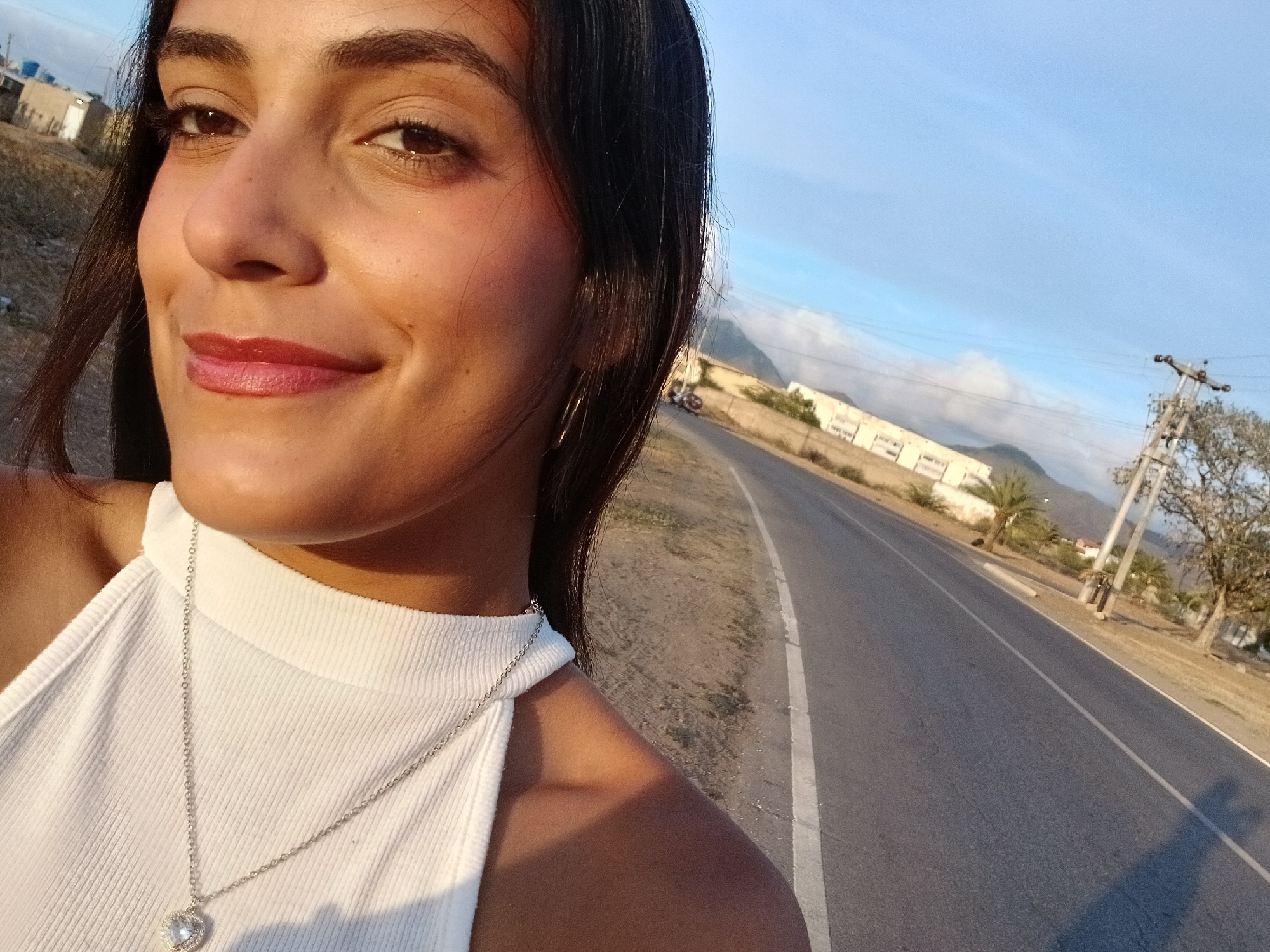 A young woman is captured in a close-up, slightly high-angle selfie-style shot, smiling gently with her head tilted. She has dark hair, dark eyes, and a warm complexion, with subtle makeup including pink lipstick and what appears to be a touch of glitter or highlight around her eyes. She is wearing a white, ribbed, high-necked sleeveless top and a silver chain necklace with a heart-shaped pendant that has a clear, sparkling stone in its center. The lighting suggests the golden hour, with warm sunlight illuminating her face and casting long shadows in the background.
The background shows an open-air scene, likely in or near Juan Griego, Venezuela. To her right and behind her, a paved road stretches into the distance, with a white line marking its edge. On the other side of the road, there's dry, light-colored terrain with some sparse vegetation, including palm trees and other trees. Several low-rise buildings with white walls are visible further back, nestled against distant, hazy mountains under a clear, light blue sky with faint wisps of clouds. A prominent utility pole with multiple cross-arms and power lines stands to the right of the road. A faint, elongated shadow, possibly of the photographer or a nearby object, is cast on the road. The overall atmosphere is pleasant and relaxed. There is no discernible text visible in the image.