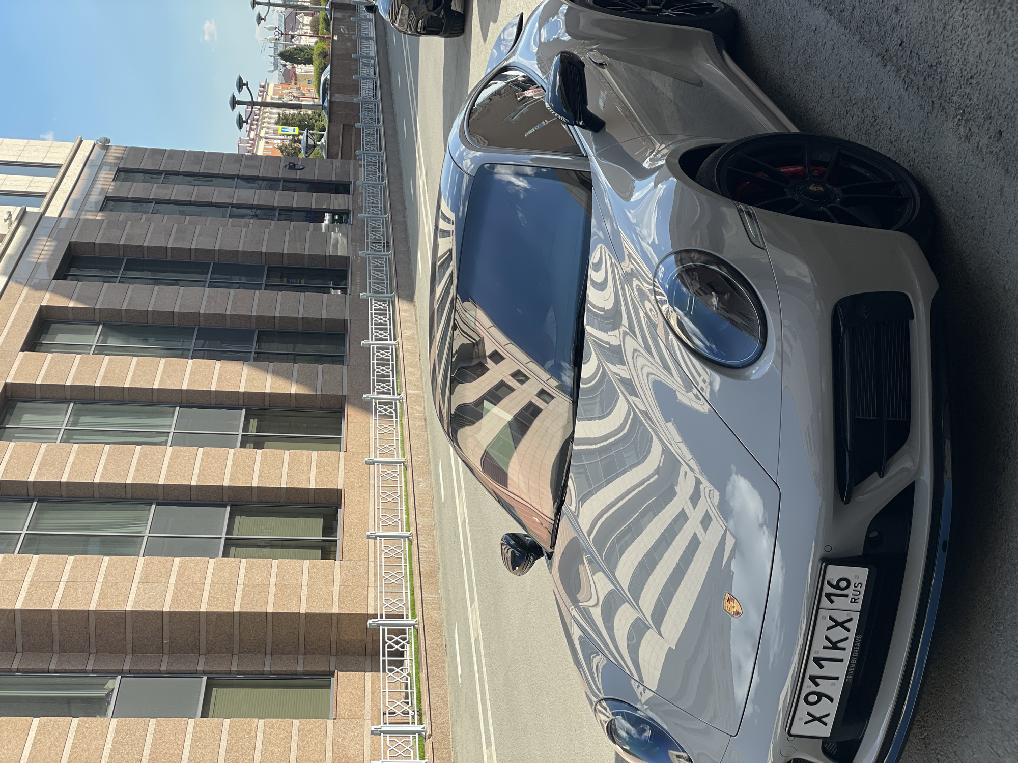 The image shows a silver Porsche 911 parked on a street next to a large, multi-story building with a façade of light brown stone tiles. The building has several rows of windows, and its reflection is visible on the Porsche's hood and windshield. A white metal fence runs along the sidewalk between the street and the building. The car has a Russian license plate "X911KX 16 RUS". The location is in an urban area, but the specific city and country are not identifiable from the image.