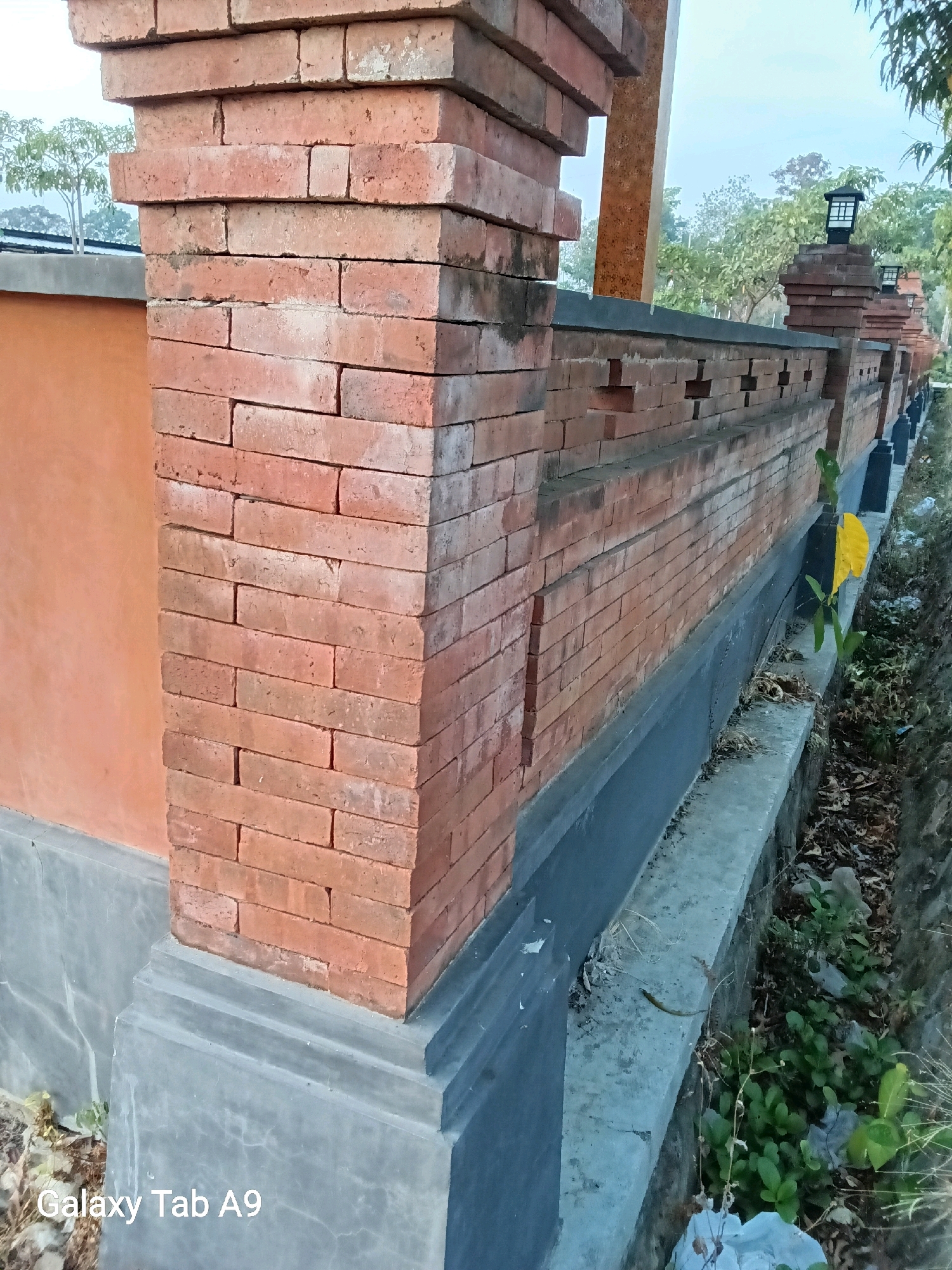 The image shows a brick fence with a textured orange wall to the left and a drainage ditch to the right. The fence is constructed of red bricks and features decorative cutouts. Several black post lights are visible along the top of the fence. The ground is covered with dry leaves and sparse vegetation. The background reveals trees and a hazy sky. In the lower left corner, the text "Galaxy Tab A9" is overlaid on the image. The scene appears to be an outdoor area, possibly a garden or a boundary wall.