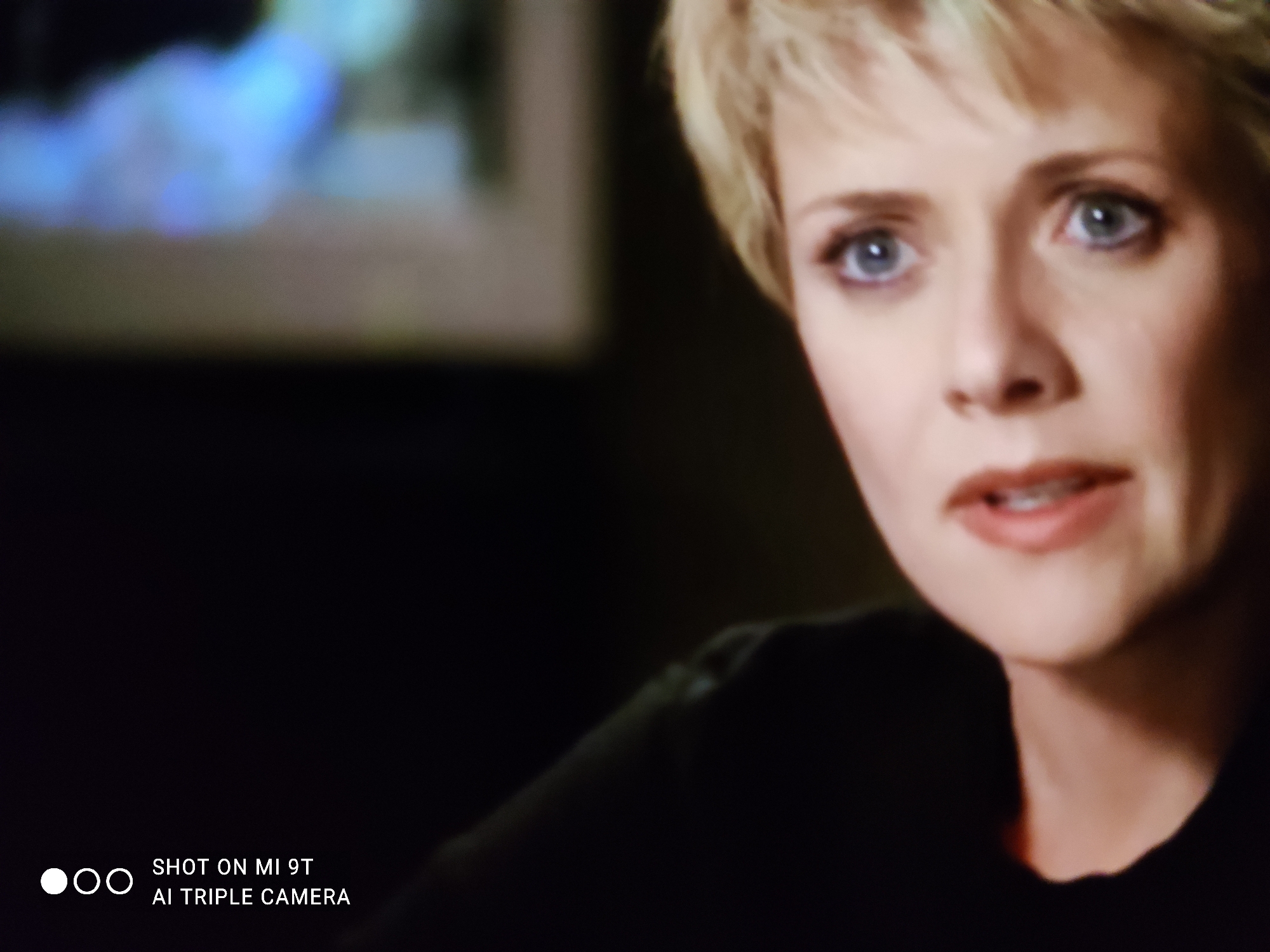 The image is a close-up shot of a woman's face, primarily focusing on her eyes and mouth. She has short, blonde hair and light-colored eyes. Her expression appears to be one of mild surprise or concern. She is wearing a dark-colored top. The background is dark and out of focus, with a hint of a blurry, light-colored object on the left side. The lower-left corner of the image contains a watermark that reads "SHOT ON MI 9T AI TRIPLE CAMERA". There is no discernible information in the image that indicates a location in Khazar Raion, Azerbaijan, or any other specific geographical area.