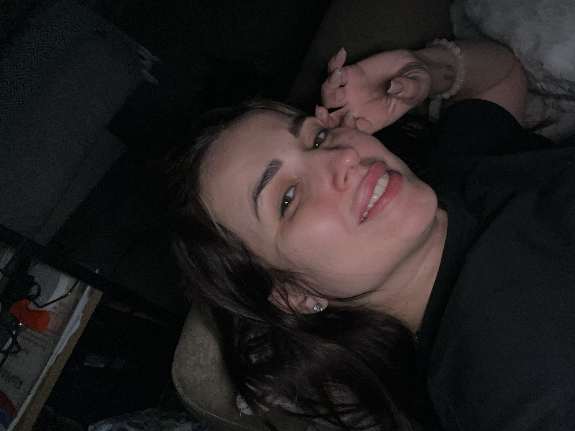 A medium shot shows a woman lying down. Her head rests on a patterned surface, possibly a cushion or pillow. She has long dark hair, fair skin, defined eyebrows, and a small stud earring. Her right hand is raised near her face, and she is wearing a bracelet on her wrist. She is smiling slightly, with her tongue visible. She wears a dark garment covering her torso. The background includes a dark sofa and some indistinct objects. The location is Unknown, Unknown.