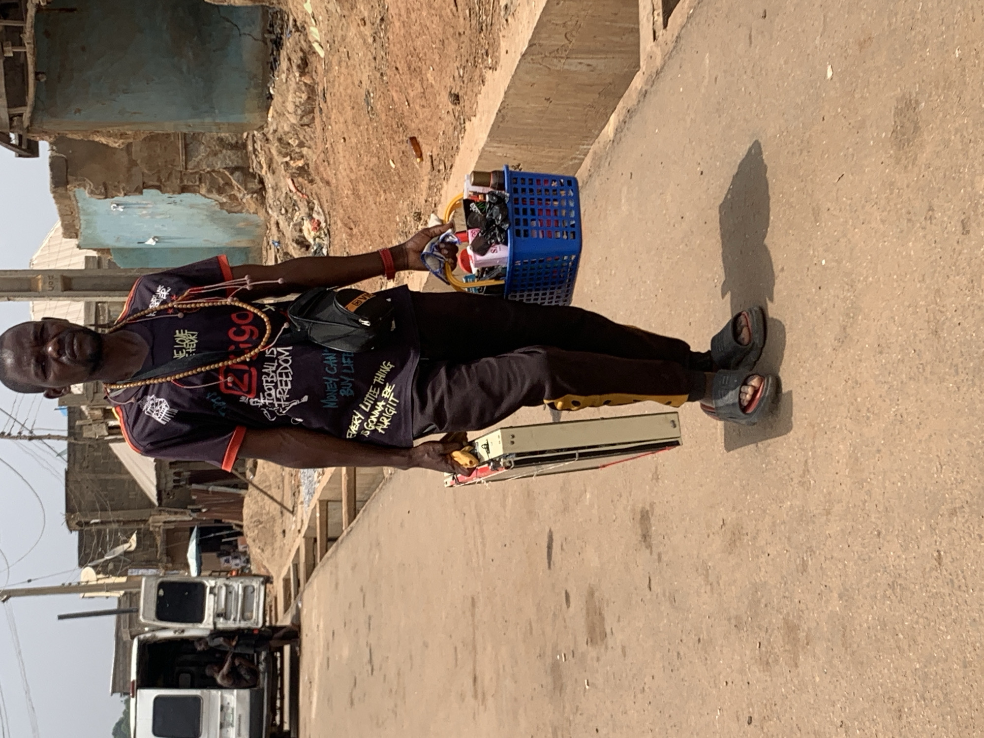 A man stands on a paved road in Akwanga, Nigeria. He faces forward, holding a blue plastic basket in his left hand and a white electric appliance in his right. He wears a dark-colored t-shirt with an orange collar and various printed words, black pants with a yellow stripe, and open-toed sandals. A shoulder bag hangs on his left side, and he has a beaded necklace.
In the background are buildings, some appearing deteriorated, and trucks parked nearby. The road is visible in the foreground, showing shadows cast by the man and objects he carries. The setting suggests an urban environment.