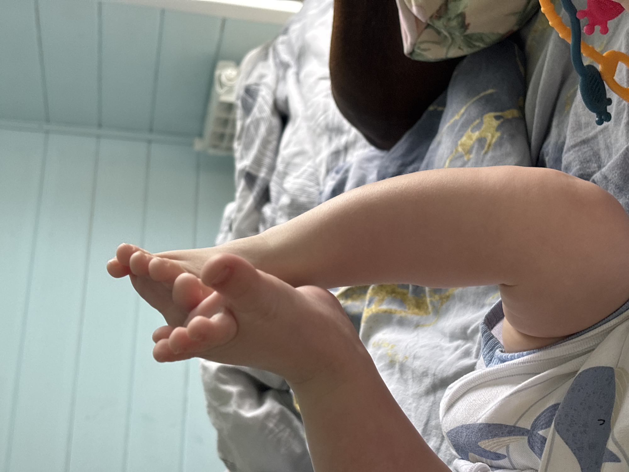 A close-up shot shows a baby lying on a bed with blue and gold patterned sheets. The baby's feet are in focus, with the toes curled slightly. The baby is wearing a white and blue pajama top with a whale design. A colorful toy hangs above the baby. The background includes a light blue wall with vertical panels and a white air conditioning unit. This image was not taken in Раздолье, Russia.