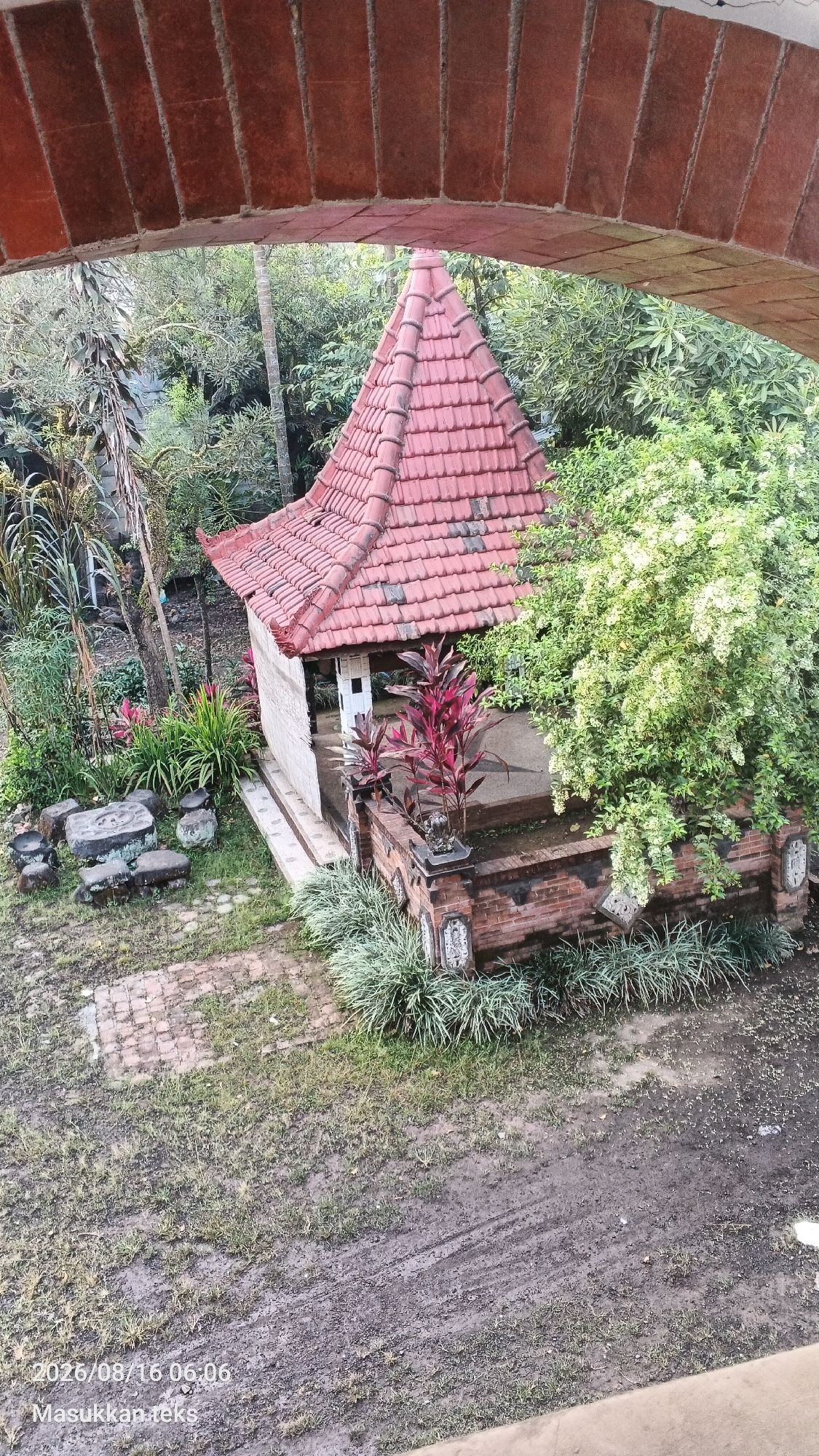 The image, taken on August 16, 2026, at 06:06, captures an outdoor scene within the city of Kendalpayak, Indonesia. A small, open-sided structure with a red-tiled, pyramidal roof is centrally located. The structure is surrounded by lush vegetation, including trees, tall grasses, and flowering plants. In front of the structure, there is a low brick wall adorned with decorative elements. Scattered around the area are various rocks and what appear to be paving stones or bricks. The ground is a mix of grass and muddy, tire-tracked earth, suggesting it might be a pathway or an area that has recently experienced vehicle activity. The overall impression is a tranquil, garden-like setting. There are no people visible in the image, and no discernible actions or interactions are taking place. The environment appears to be a humid, tropical climate, with the greenery indicating a high level of rainfall or irrigation.