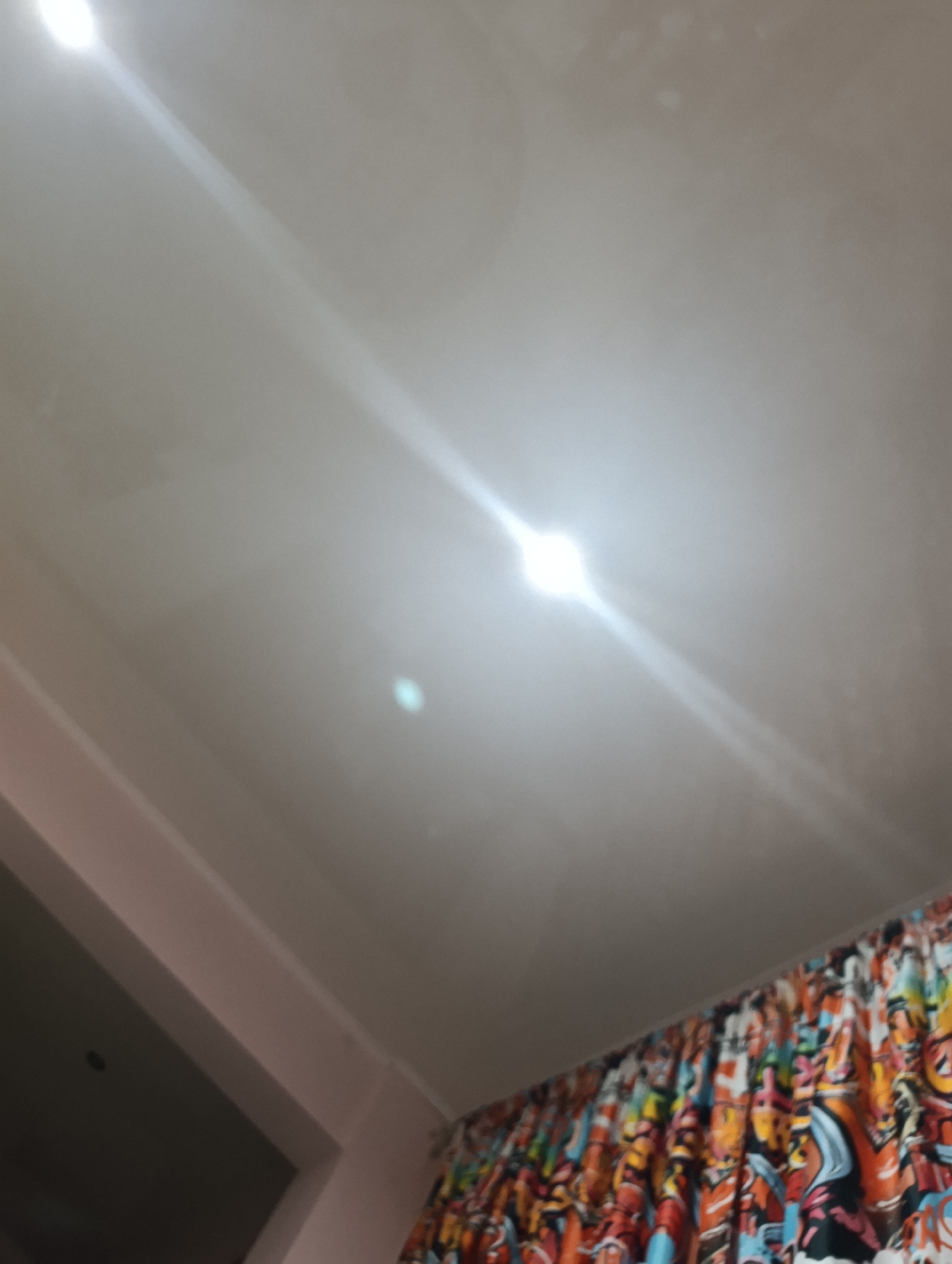 The image shows a ceiling with bright light reflections. On the right side of the image, there is a colorful curtain with a graffiti-like pattern. The lower left corner reveals a part of a wall and what appears to be a doorway. There are no people or discernible actions taking place. The environment suggests an indoor setting. The provided crops offer closer views of the ceiling with light flares and the detailed pattern of the curtain. There is no information within the image to suggest a location in Astrakhan, Russia.