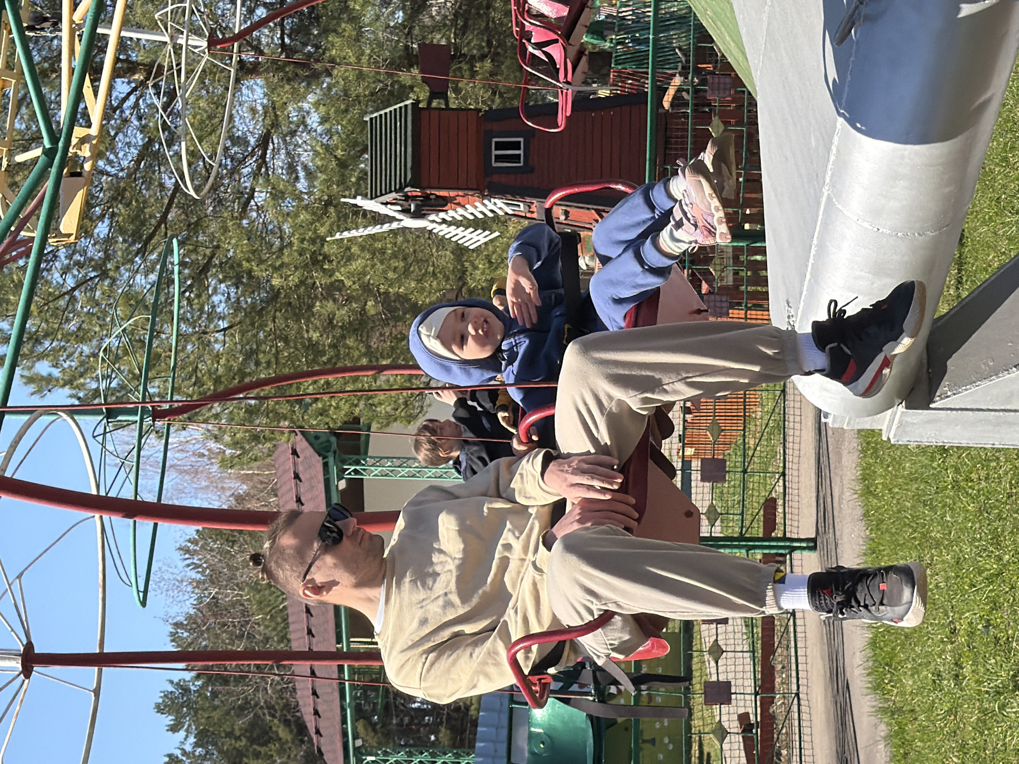 The image depicts an adult male and a young child riding a carousel at an outdoor location. The man is wearing sunglasses and a beige sweatsuit. The child is dressed in a blue hooded jacket and pants. Both are seated in red seats suspended from the carousel structure. The carousel is constructed of metal, with red, green, and yellow support bars. The background includes trees and a small, red wooden structure resembling a miniature house or cabin. Green grass is visible along the bottom and right edges of the image. The image was taken in the city of Chaikovsky, Russia.