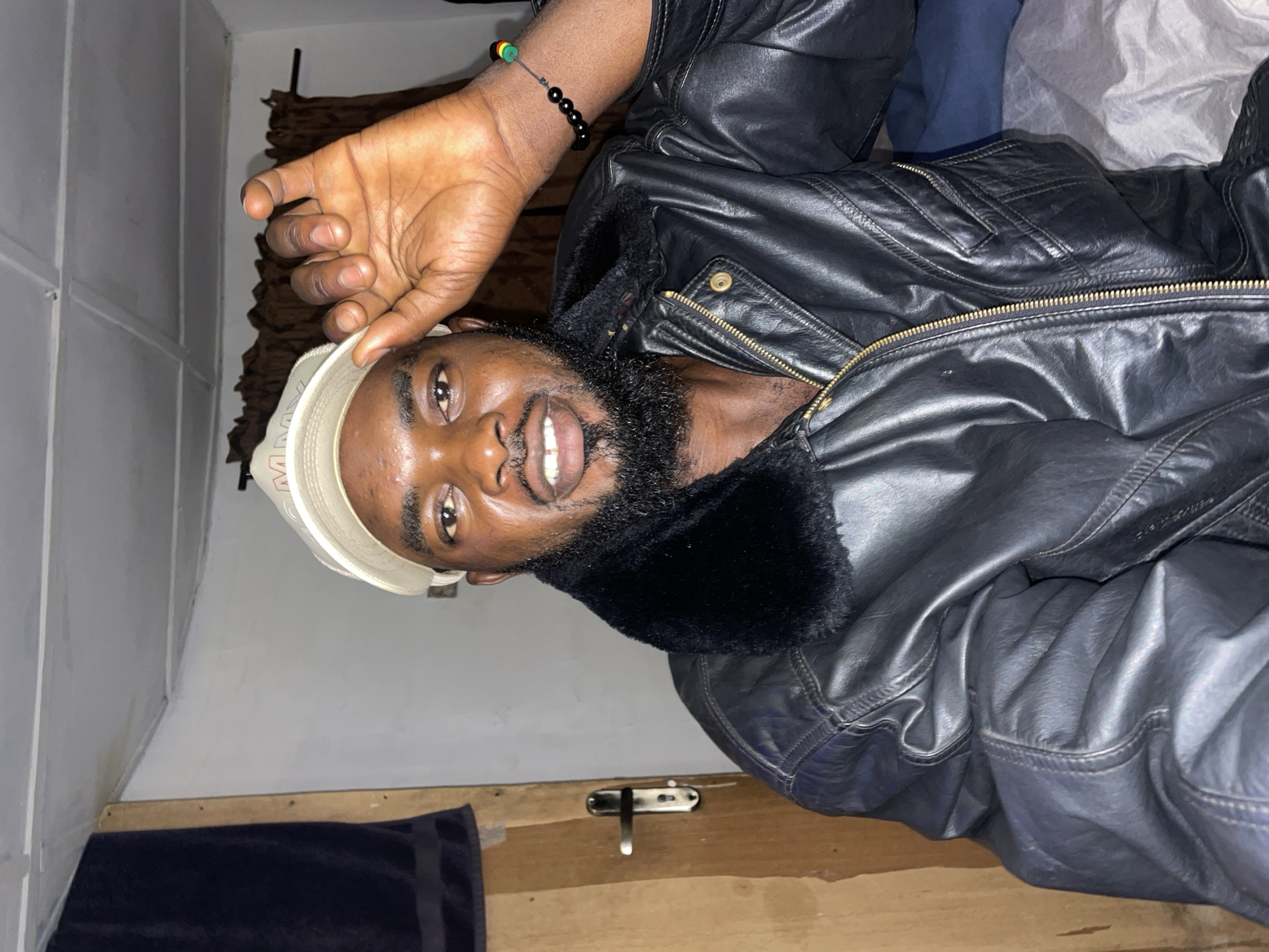 A man with a beard and a light-colored baseball cap is lying down, wearing a black leather jacket with a fur collar. He is smiling and holding his hand up near his face. The background includes a white wall with paneling, a brown curtain or fabric, and a wooden surface with a metallic latch. No specific location is discernible from the image to determine if it is in Jos, Nigeria.