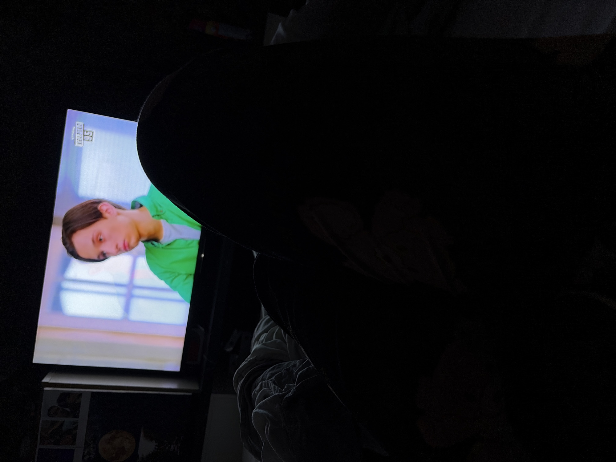 The image shows a person lying in bed at night, watching television. The person is mostly in shadow, with only their legs and torso visible. The television screen is on and shows a young man in a green shirt. There are some framed pictures on the wall to the left of the television. The room is dark, and the scene is dimly lit by the television screen. This appears to be a personal bedroom within a residential setting in Amstelveen, Netherlands.