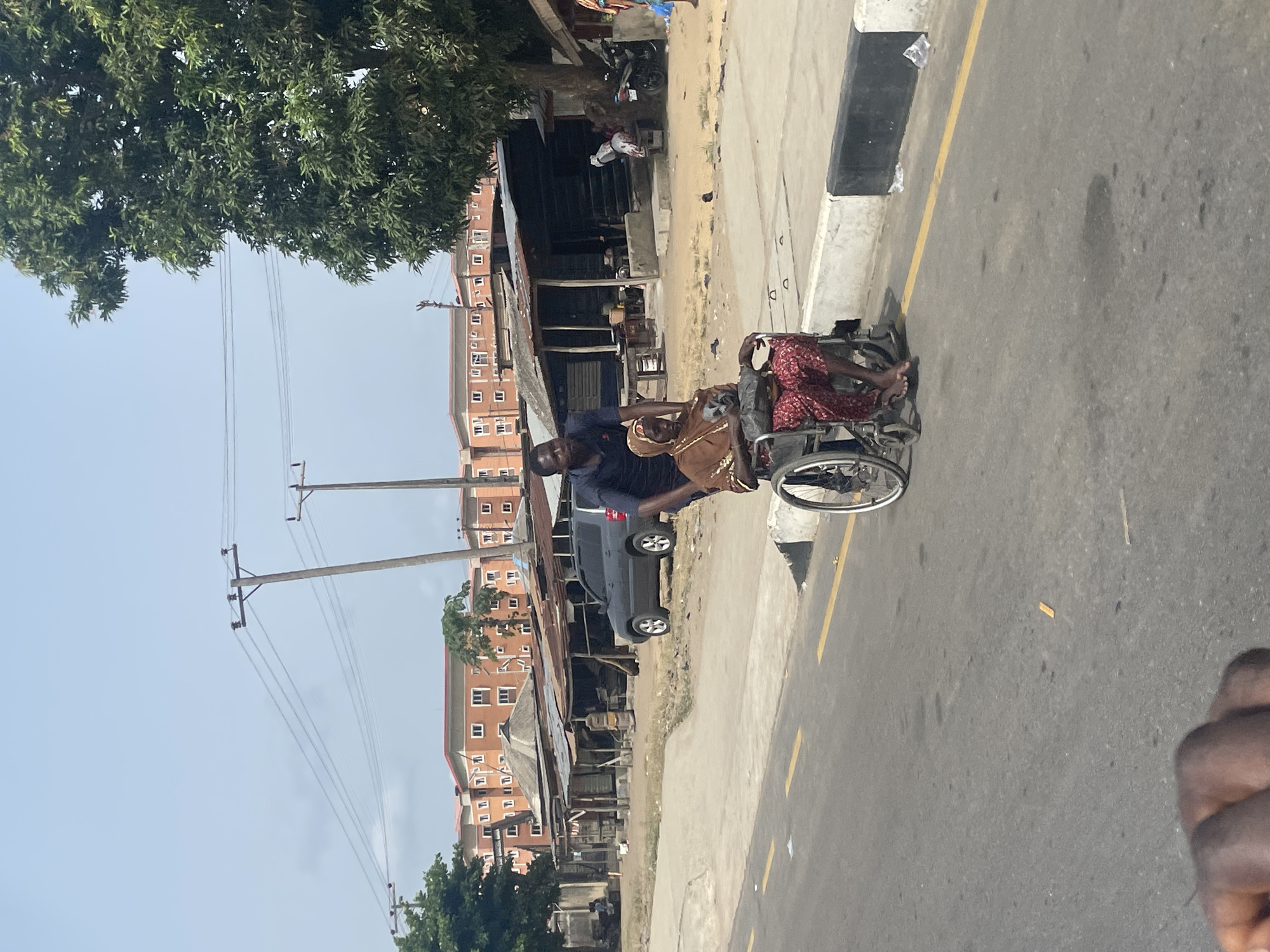 The image shows a man pushing a woman in a wheelchair on a paved road in Ajara, Nigeria. The woman wears a head covering and red patterned dress and her feet are bare, while the man wears a dark blue shirt. The location appears to be near a roadside market or commercial area, with buildings, shops, and parked vehicles visible in the background. Utility poles and trees line the other side of the road. A roadside curb is visible adjacent to the paved road. The sky is light blue.
