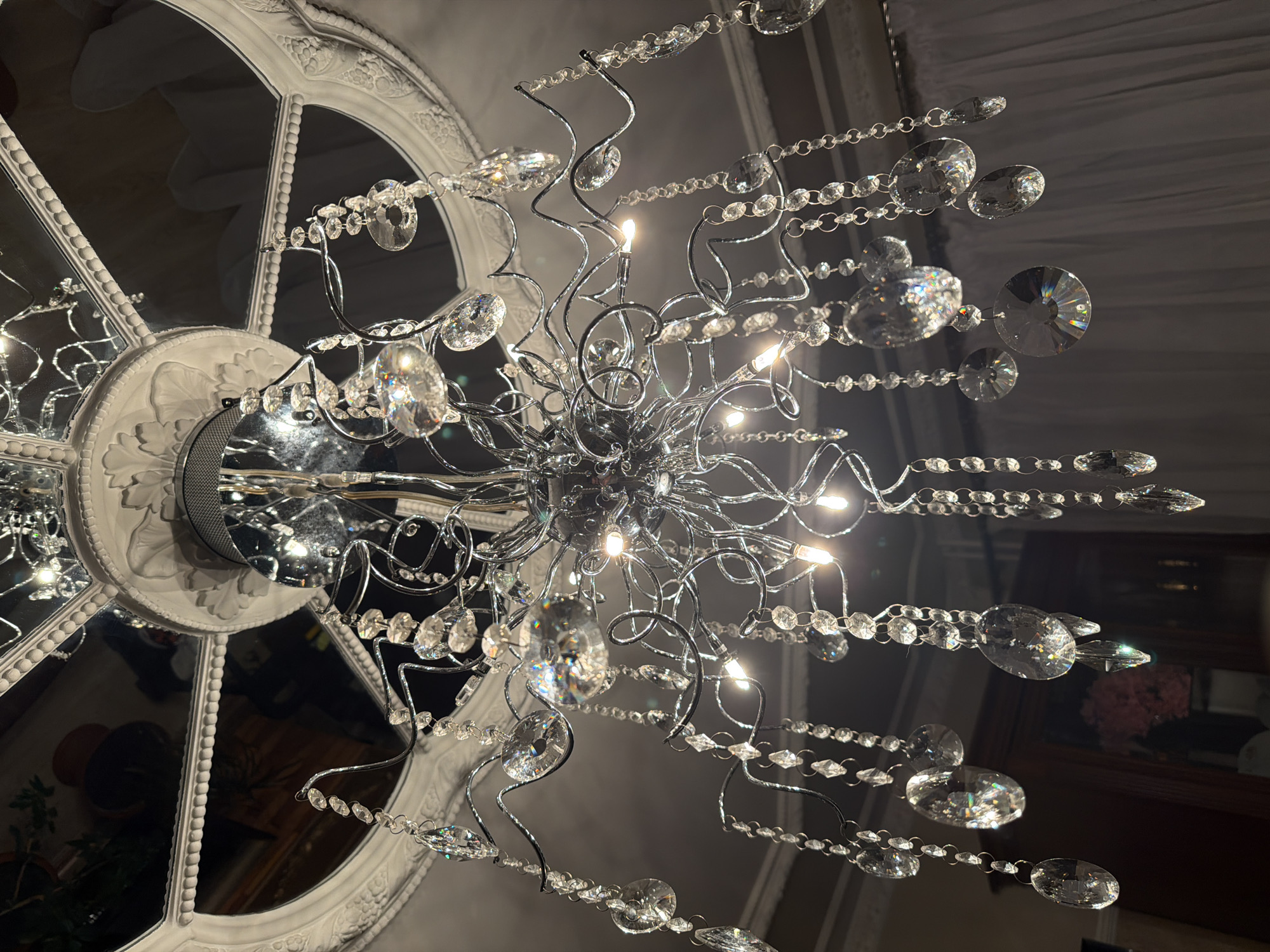 A low-angle, indoor photograph shows a chandelier with strings of small, round, and disk-shaped clear crystals suspended from a central fixture of shiny silver metal in front of a large, round, white-painted window frame. The chandelier lights are illuminated, casting light in the space. The window frame is attached to the ceiling. The chandelier hangs from the ceiling within the room. There is no indication of the image's location in the city of Krasnoyarsk, Russia.