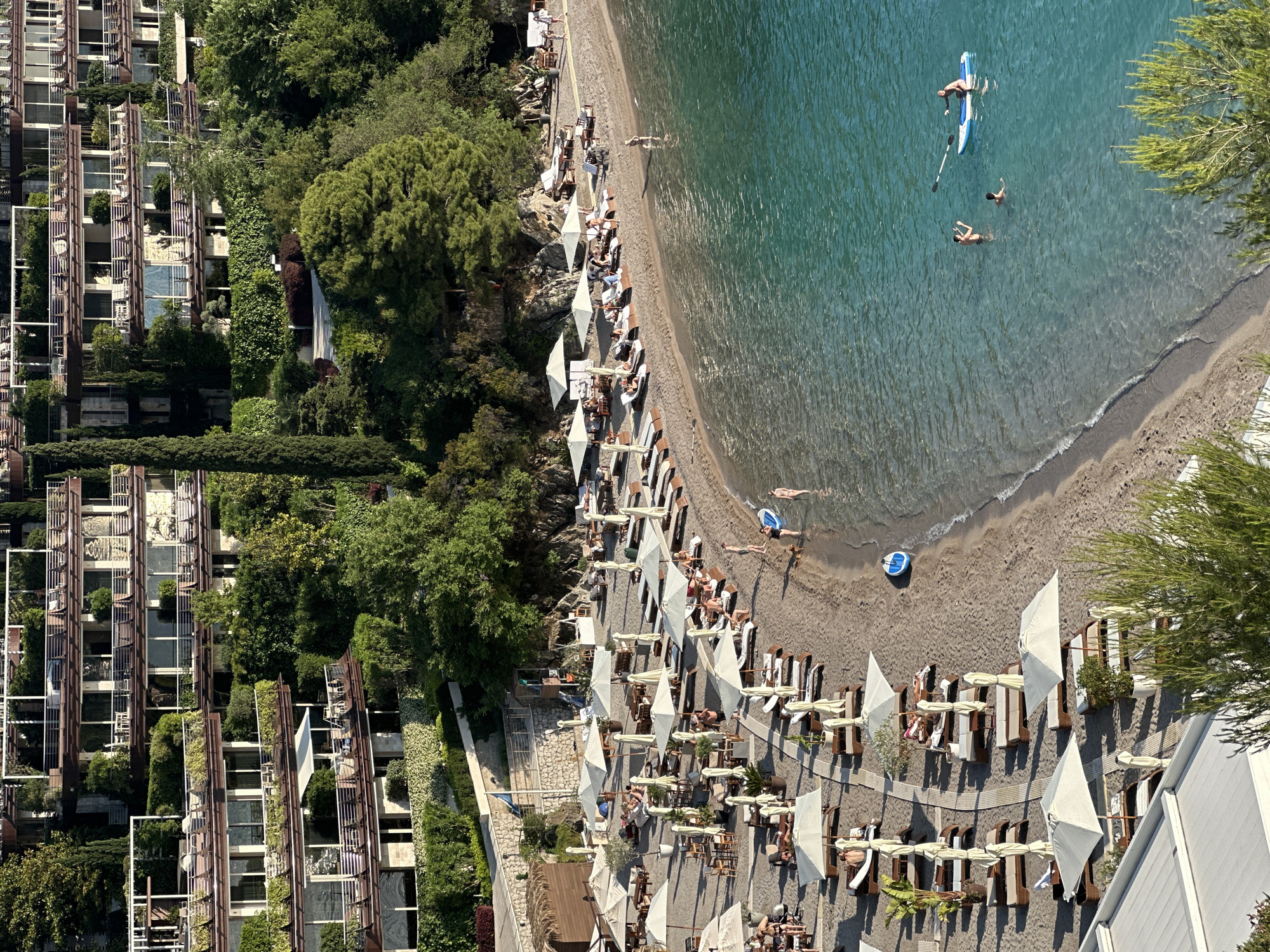 The image depicts a beach scene in Budva, Montenegro. A series of tall, modern buildings, heavily vegetated, are located adjacent to the beach. Rows of beach chairs and white umbrellas line the sandy shore. People are lounging on the beach, some are swimming, and one person is on a paddleboard in the clear, blue water. The overall view is from an elevated perspective, capturing both the architecture and the beach activities.