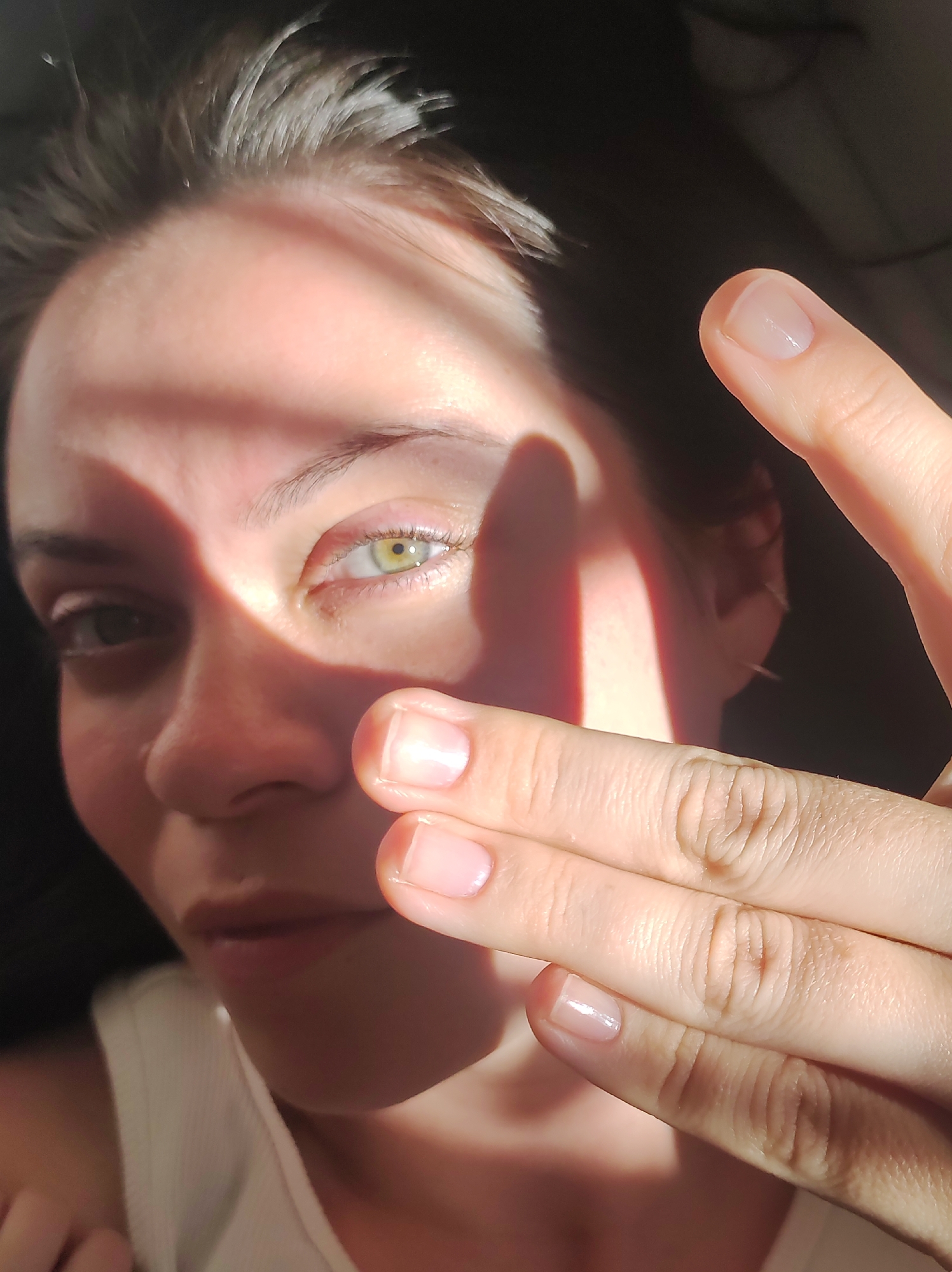 A close-up selfie of a woman with fair skin. Her face is partially obscured by shadow. Her left eye is green. She is holding her hand up, obscuring parts of her face. Her fingernails are filed short and covered in a clear coat. The location is indeterminable.