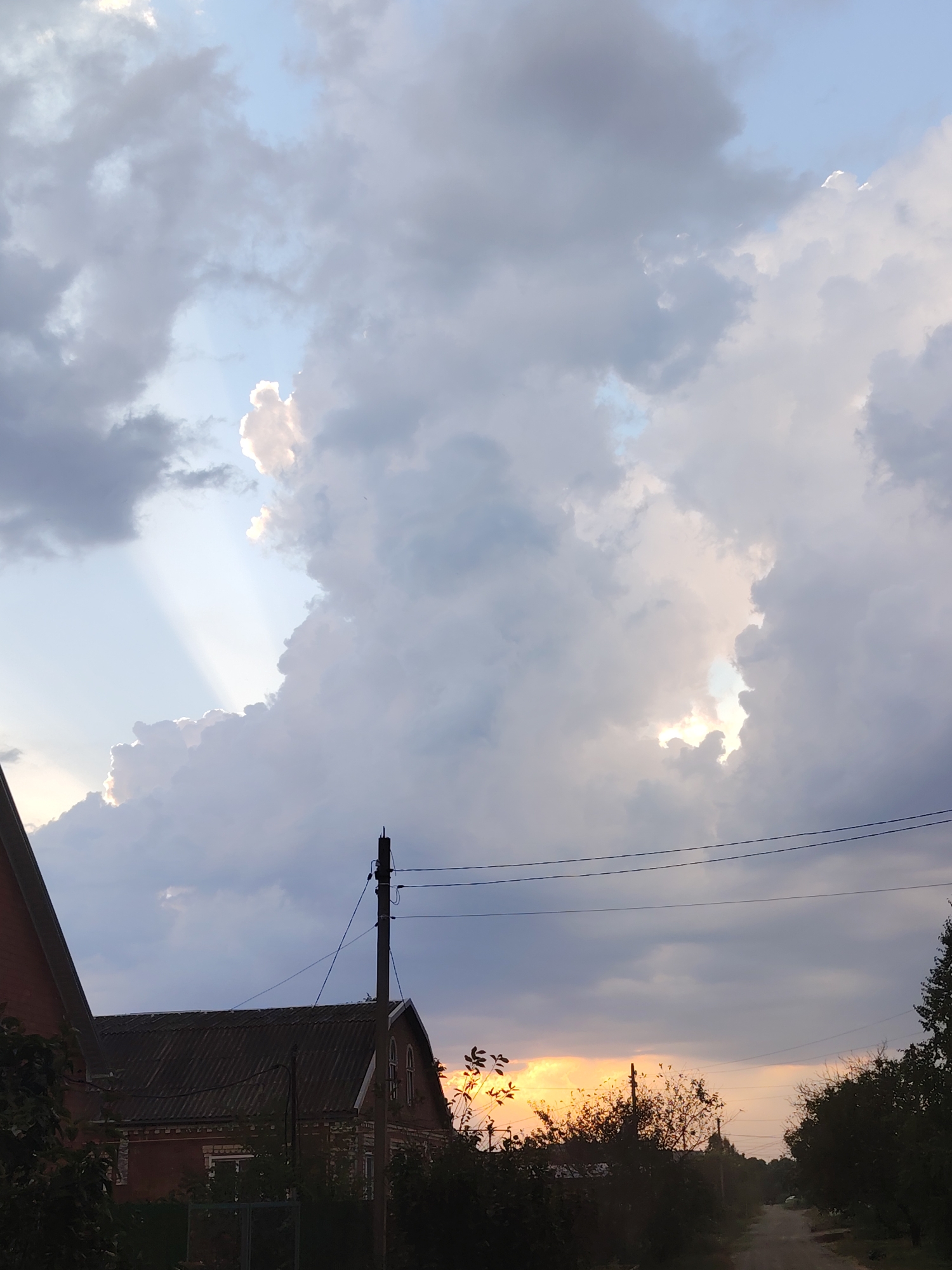 This is a photograph taken from a low angle, looking up at a dramatic sky above a rural village. The sky is filled with large, billowing clouds, some of which are dark and heavy, while others are illuminated by the sun peeking through. Shafts of light are visible breaking through the clouds, creating a dramatic effect. In the foreground, the roofs of houses are visible, along with trees and a power pole with wires. A dirt road leads into the distance. The overall impression is one of a stormy but beautiful sunset. The location is the village of Volnoye, Russia.