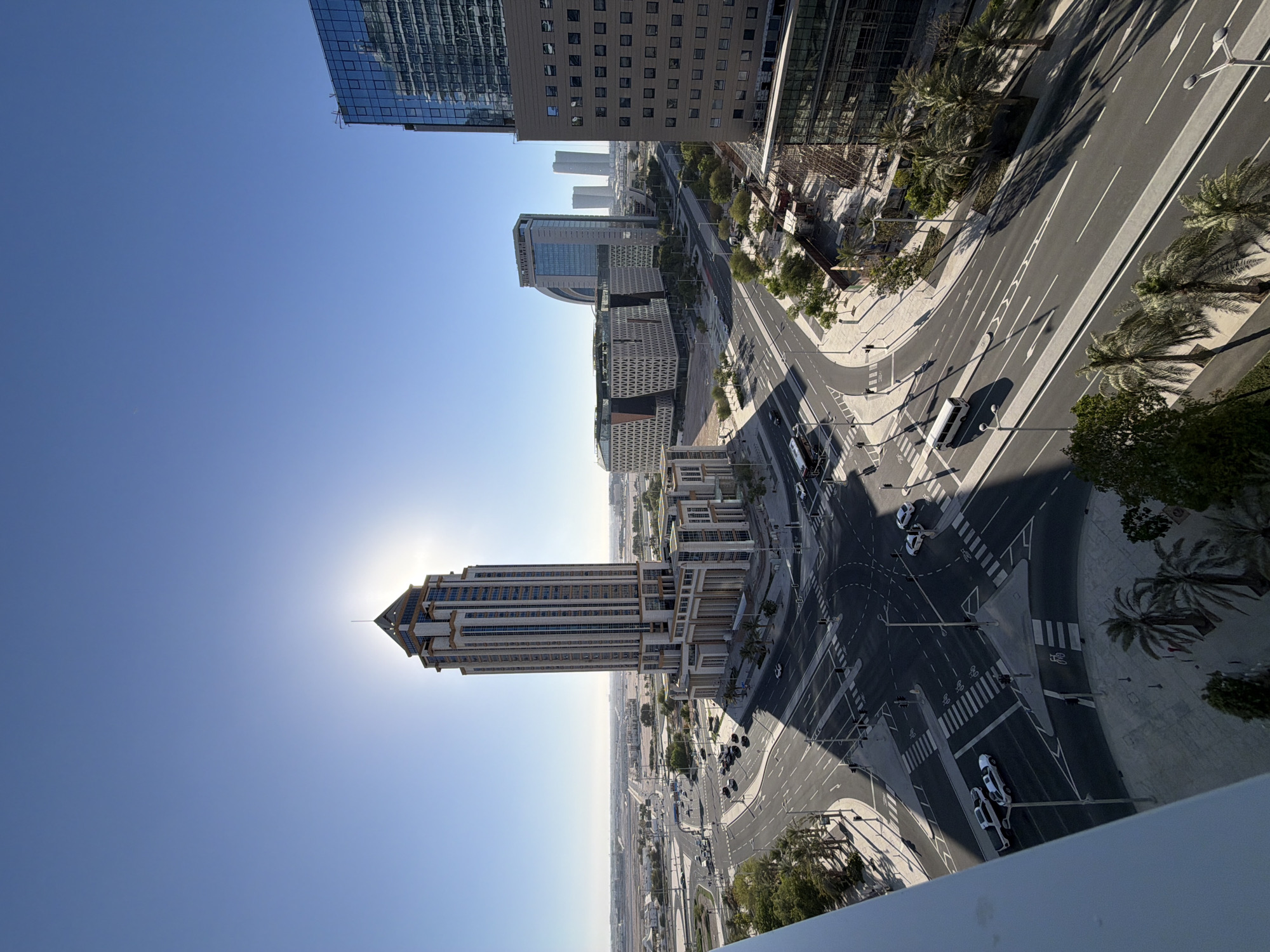 A high-angle, eye-level, wide shot captures a modern city intersection under a clear blue sky. The sun is high and bright, positioned behind the peak of a tall, multi-story building on the left side of the frame, creating a sun flare effect and casting long shadows. The building has a distinct pyramid-like top with a small spire.
The intersection itself is a complex network of asphalt roads with white lane markings. Pedestrian crosswalks are clearly visible. Several vehicles are navigating the intersection: a white pickup truck and two white sedans are stopped at a crosswalk, a white van is traveling on the road, and a few other cars are visible in the distance.
Surrounding the intersection are various modern buildings. To the left of the tall building, there are lower-rise structures and a vast expanse of what appears to be undeveloped land or a distant cityscape. In the center, a large, curved building with a patterned facade stands out, adjacent to another modern building with a glass facade. To the right, a tall, glass-clad building reflects the sky and surrounding structures. Construction scaffolding is visible near the base of this building.
Palm trees and other scattered greenery dot the landscape around the roads and buildings, suggesting a warm climate. The overall scene depicts a well-developed urban environment with active traffic flow.