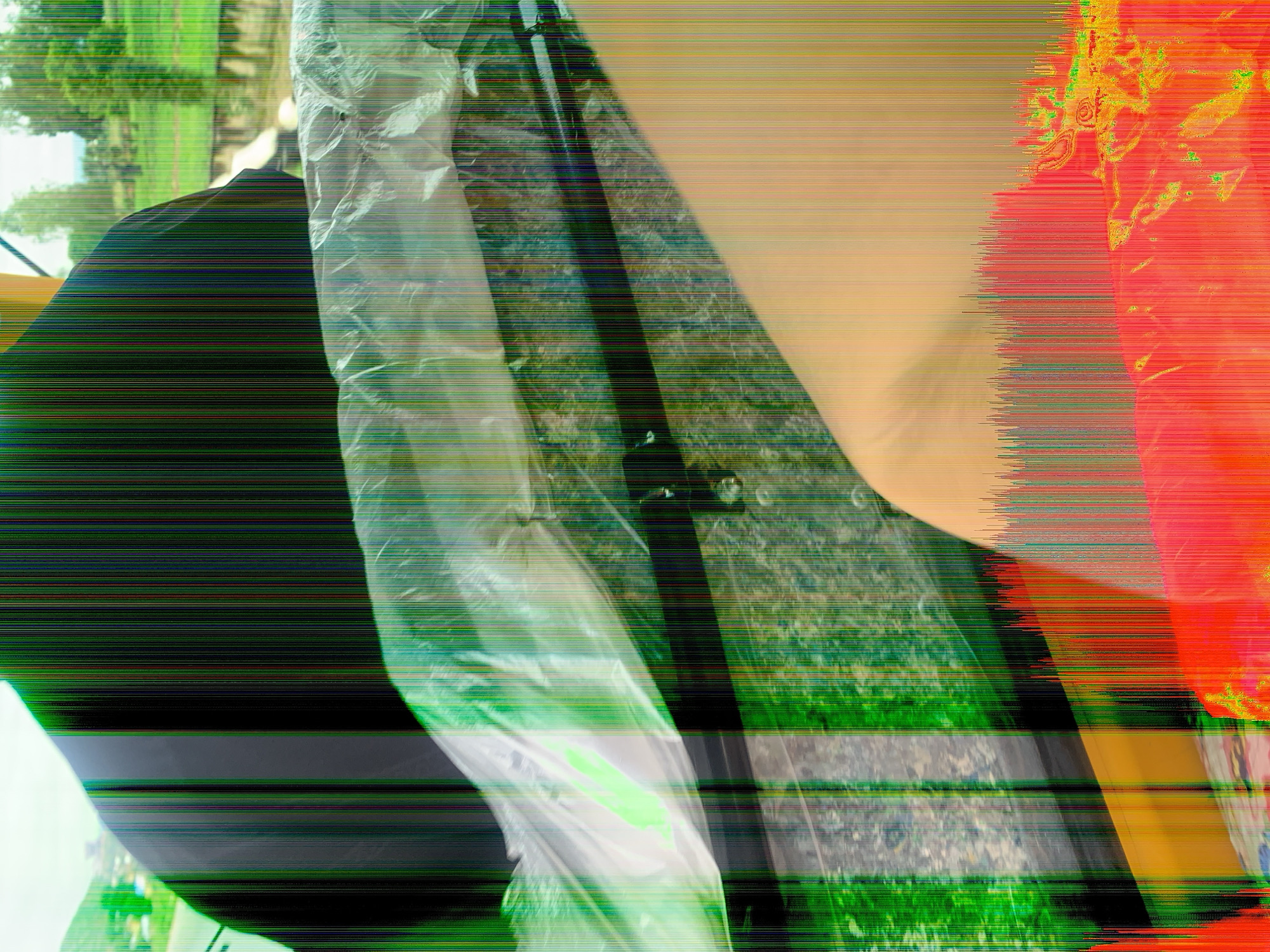 The image displays a distorted view of a covered object with vertical streaks of color throughout. In the upper left, a green, hilly landscape with scattered buildings is visible, suggesting an outdoor setting in Jalingo, Nigeria. The primary focus is on a large, dark, curved object, possibly a vehicle or piece of equipment, shrouded in clear plastic sheeting. The sheeting reflects light and shows some texture beneath. Below this, a textured, mottled surface, perhaps stone or concrete, is visible. The lower portion of the image is dominated by bright, abstract shapes in orange and red, with a dense scattering of vertical red, green, and blue lines, indicative of digital artifacting or visual interference.