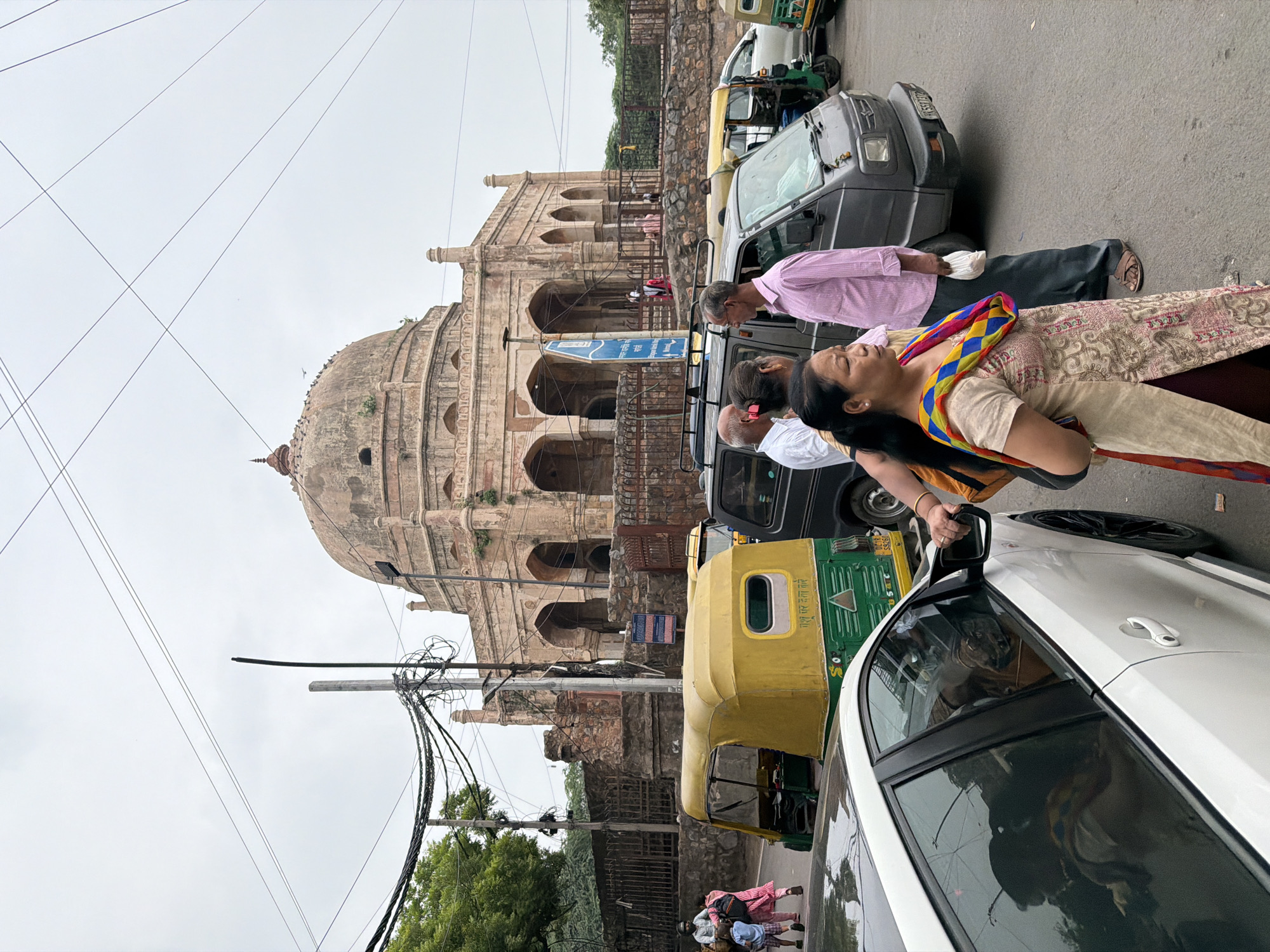 The image captures a street scene in Delhi, India, with a prominent historical monument in the background. The monument features a large dome and arched openings, suggesting it is a tomb or mosque. In the foreground, a white car is parked, and a woman in traditional Indian attire with a colorful scarf is standing next to it. Several auto-rickshaws, a common mode of transport in Delhi, are also visible, along with other vehicles and pedestrians. The sky is overcast, indicating a cloudy day. The area appears to be a busy urban environment, with the juxtaposition of modern vehicles and ancient architecture.