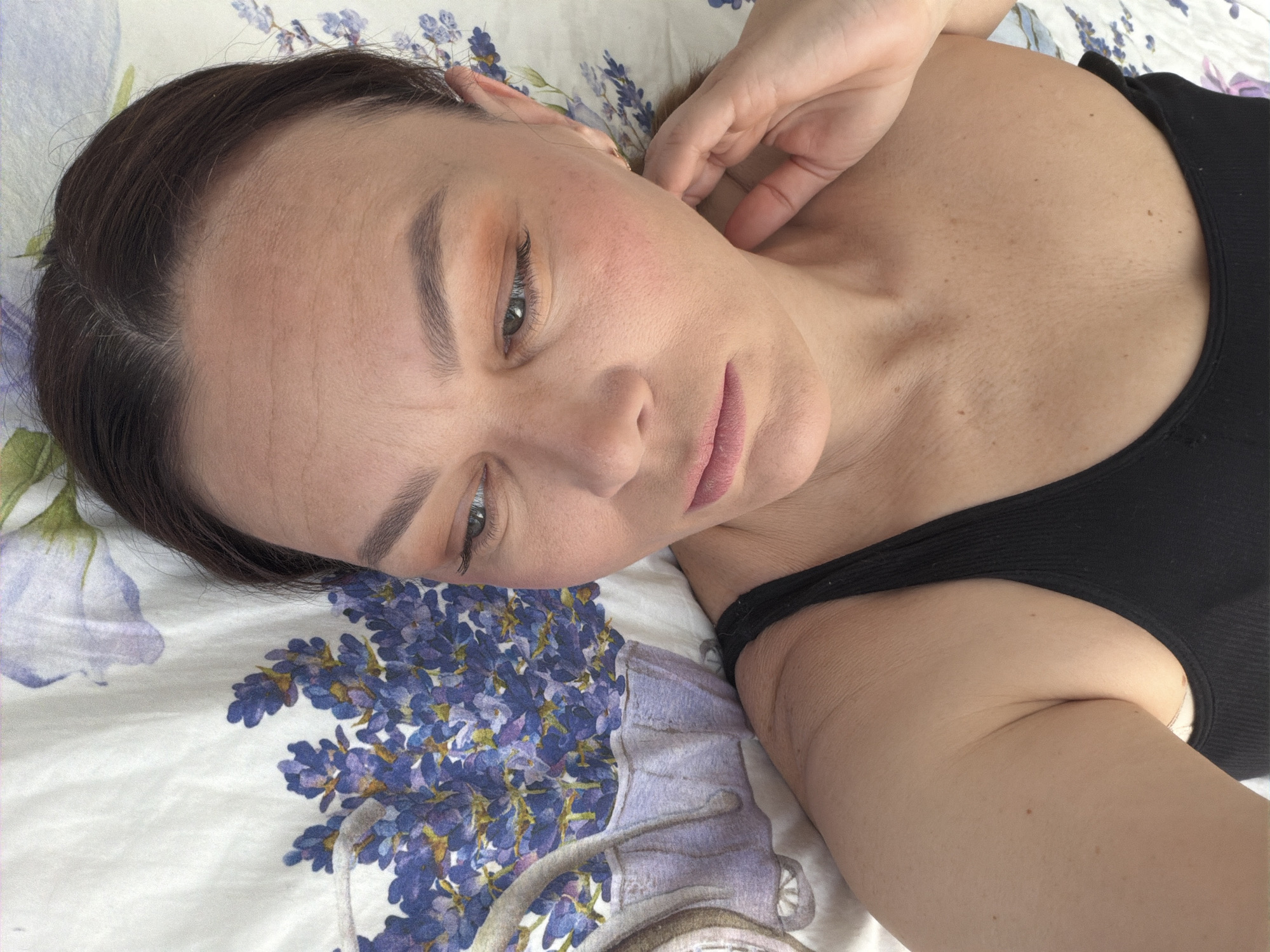 A fair-skinned woman lies on a bed with lavender-patterned bedding. She wears a black tank top and has dark hair pulled back. Her right hand is raised near her neck. The location is unknown and cannot be determined from the image.