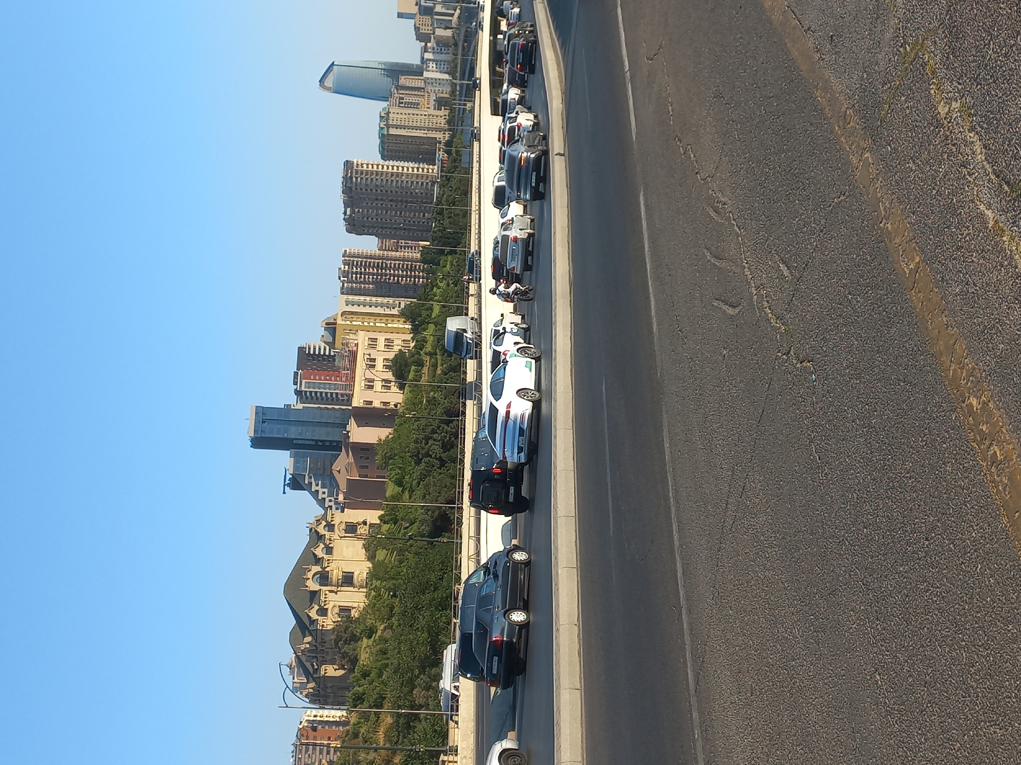 The image depicts a busy urban scene in Baku, Azerbaijan, under a clear blue sky. The foreground shows a multi-lane road with various vehicles in traffic. A grey sedan and a white Porsche are prominently featured. In the background, a cityscape unfolds with a mix of architectural styles. A grand, ornate building with a dark green roof stands on a hill, flanked by trees and other structures. Further in the distance, modern high-rise buildings, including a distinctive glass skyscraper with a curved top, dominate the skyline. The overall impression is one of a developed city with both historical and contemporary elements.