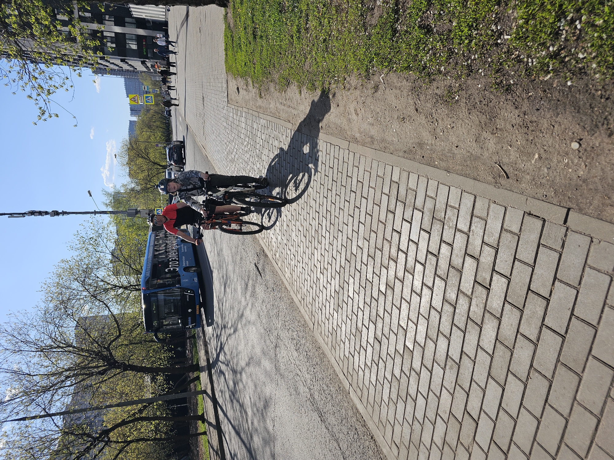 A sidewalk in Khimki, Russia, is paved with rectangular gray bricks and is adjacent to a patch of grass. Two people are riding bicycles on the sidewalk, casting shadows on the brick surface. A blue bus is visible in the background. Pedestrians and cars are on the road. The weather is sunny with trees and buildings along the road. A blue traffic sign is visible in the distance.