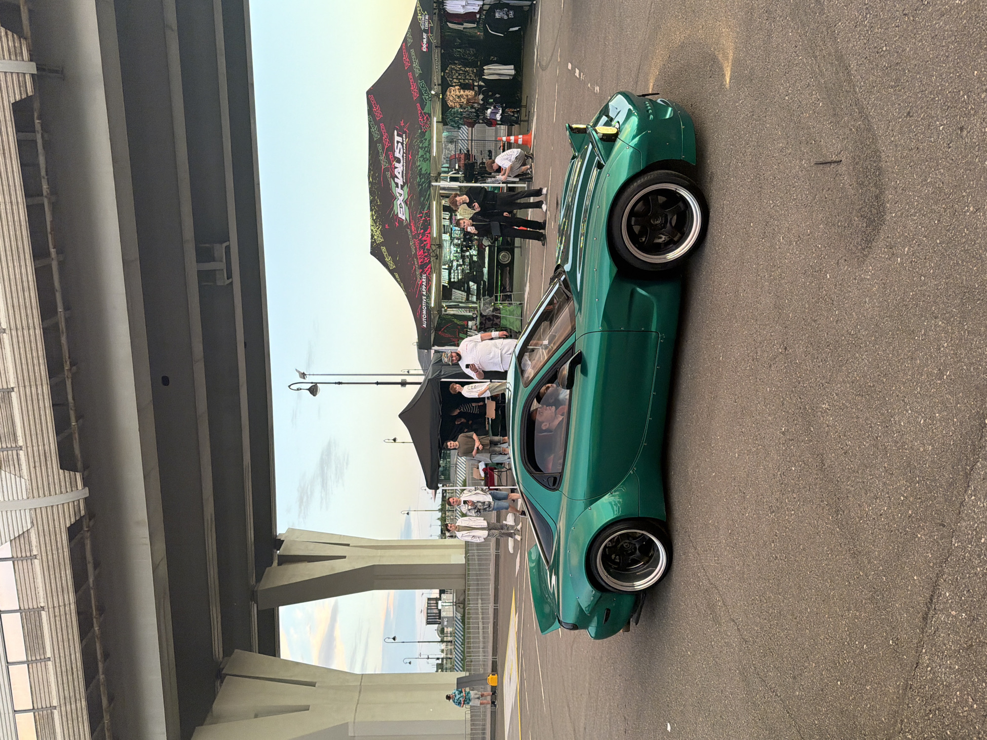 A green sports car is parked on a paved surface, likely an outdoor event. The car is a wide-body Mazda RX-7 FD. In the background, there are several tents, including one branded "EXHAUST AUTOMOTIVE APPAREL." A group of people are standing near the tents, some looking at their phones, others interacting. A large concrete structure, possibly a bridge or overpass, looms over the scene. The sky is a pale blue with some clouds. There are streetlights visible in the background. The car has custom wheels and a large rear spoiler.