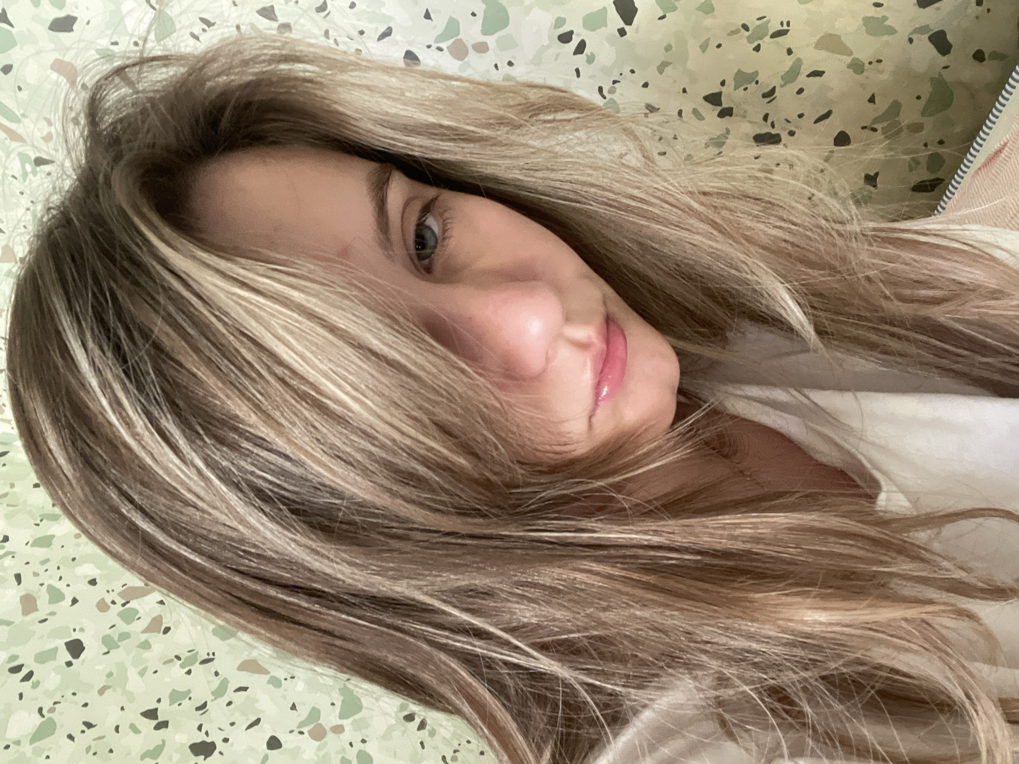 A young woman with blonde hair and blue eyes looks forward with a slightly melancholic expression, her hair falling across her face. She is wearing a white shirt and is positioned in front of a wall with a light green, speckled pattern. The lighting is soft, and the image appears to be a selfie taken indoors. The exact location in the city of Unknown, Unknown is not determinable from the visual information provided.