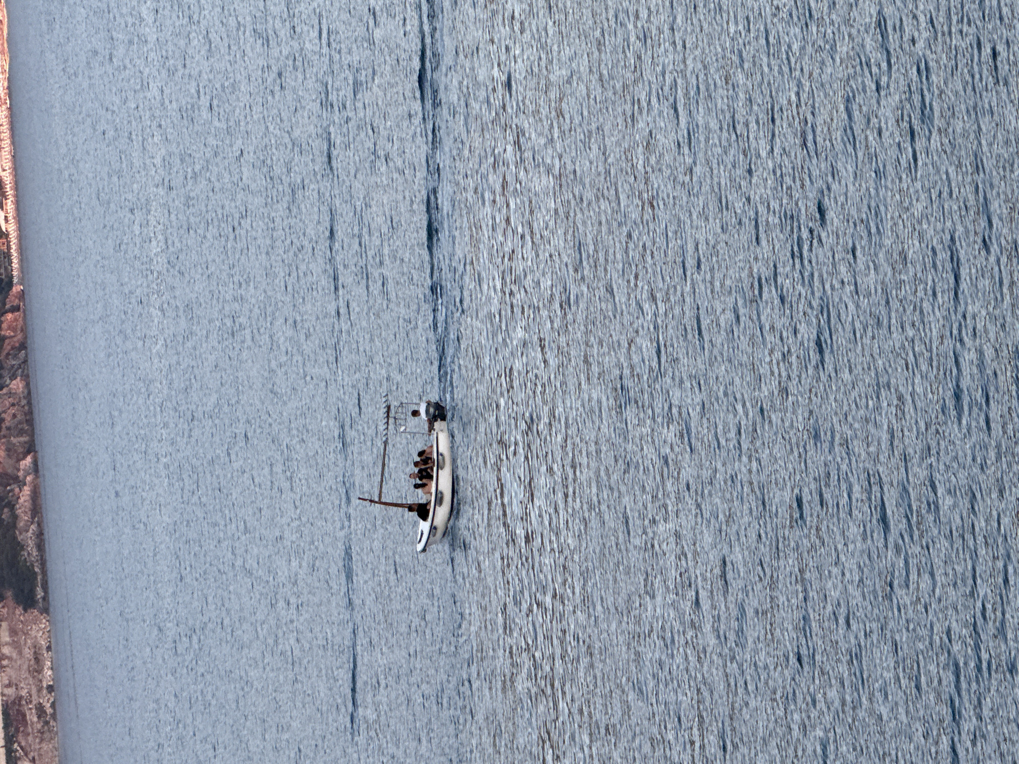 A small, white sailboat with a dark hull is sailing on a calm, blue body of water. The boat is occupied by several people, their forms clustered together. The water is textured with ripples and slight chop, creating a streaky pattern. On the left side of the image, a rocky, tree-lined shore is visible, suggesting a mountainous or hilly landscape. The scene appears to be set in a natural, outdoor environment. The lighting suggests it could be daytime, with no strong shadows indicating early morning or late afternoon. The overall impression is one of a peaceful excursion on the water, possibly in a scenic location like Boreti, Montenegro.