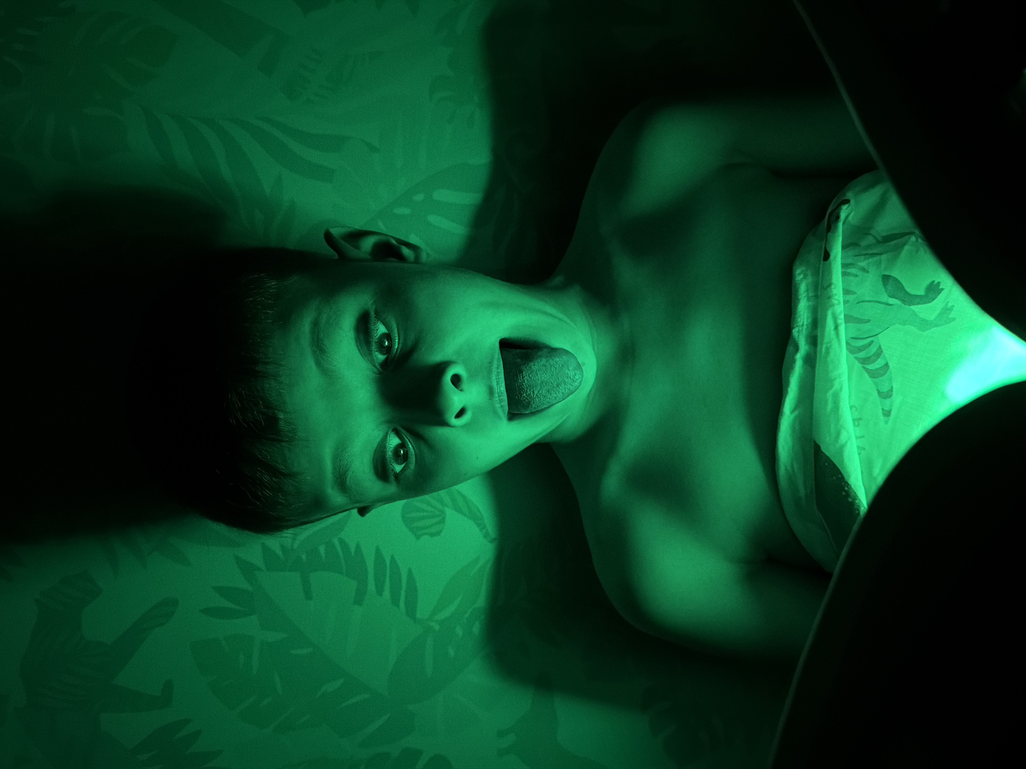 A child lies on a patterned surface, possibly a bed or sofa, with a green light illuminating the scene. The child is sticking out their tongue. A sheet or blanket with a print is partially covering the child. A dark object is obscuring the right side of the image. This scene appears to be indoors, likely in a residential setting. The provided information lacks any indication of this photo being taken in the city of Votkinsk, Russia.
