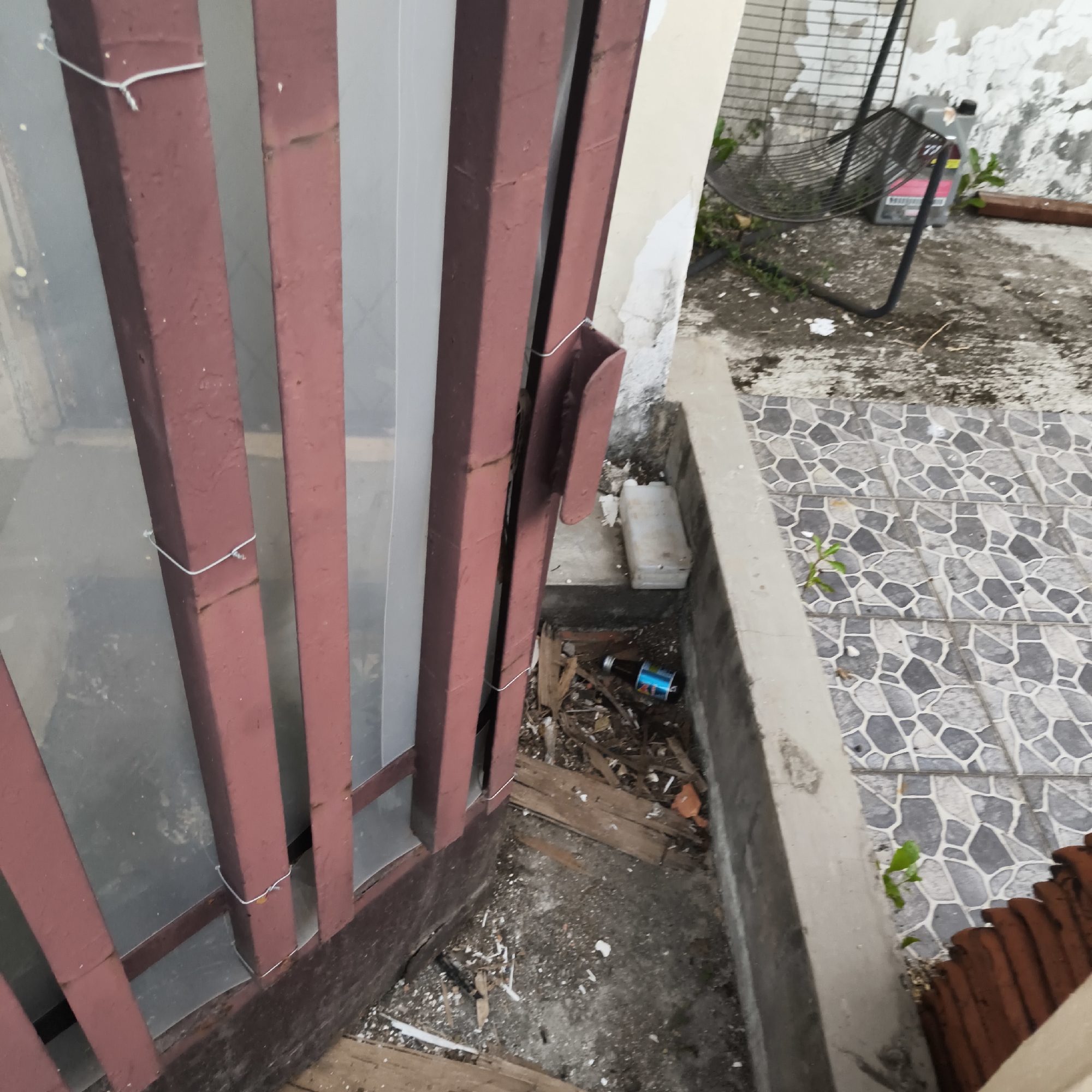 The image depicts a close-up view of an outdoor area with a fence or gate made of dark red metal bars. The ground is a mix of concrete and tiled surfaces. Debris, including wood fragments and what appears to be a discarded bottle, is scattered on the ground near the fence. In the background, part of a building with a textured, light-colored wall is visible, along with a black metal fan and some greenery. The scene suggests an unkempt or neglected outdoor space, possibly a backyard or alleyway in an urban environment. The presence of a tiled walkway and a building suggests a residential or commercial area. The lighting suggests it is daytime. The specific location in South Jakarta, Indonesia, is not discernible from the visual information provided in the image.