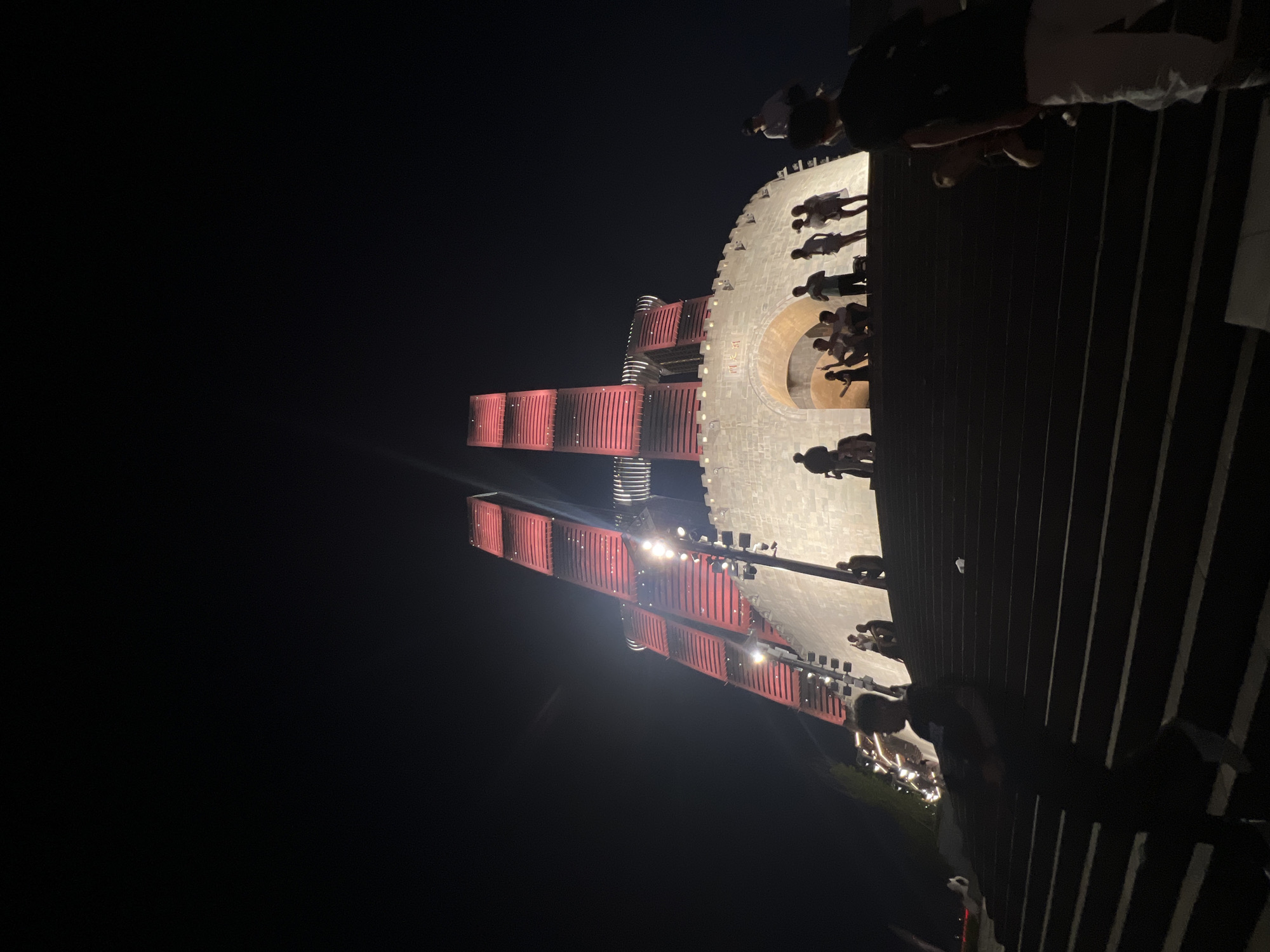 In the city of Nan'an District, China, at night, a large stone structure resembling ancient fortifications with a grand archway is illuminated. Behind it, two towering, modern architectural structures, lit with bright red lights, ascend into the dark sky. Several individuals are visible, some standing on the lower steps leading up to the stone structure, while others are gathered at the top, near the archway. One person in the foreground is silhouetted against the stairs. The overall scene captures a blend of historical and contemporary architecture illuminated for nighttime viewing.