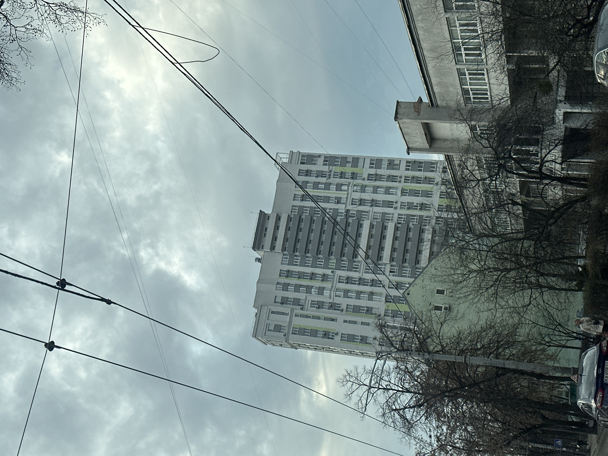 The image shows a cityscape in Kyiv, Ukraine, under a cloudy sky. In the foreground, power lines crisscross the view. A modern high-rise building with multiple floors and numerous windows is visible in the center of the frame. To the right, an older, shorter building with visible wear and tear stands among bare trees. Further to the right and partially obscured by trees, a car is parked on the side of the street. There are at least two pedestrians walking on the sidewalk to the right side of the frame. The scene suggests a blend of modern and older architecture within an urban setting.