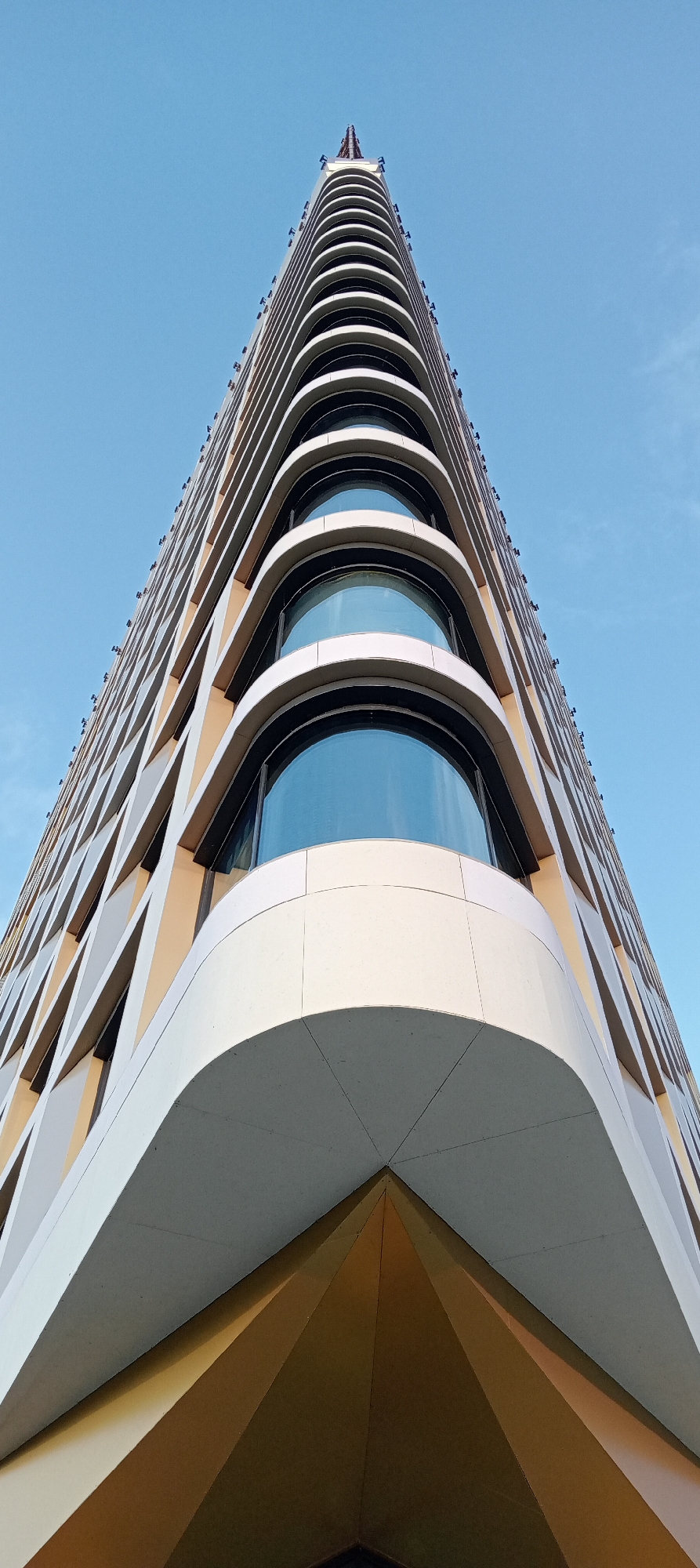 The image shows a low-angle, worm's-eye view of a modern, triangular-shaped skyscraper with a distinct stepped facade and curved windows. The building's exterior is a mix of light-colored material and gold accents, with a lattice-like pattern on some sections. The sky above is clear and bright blue. The skyscraper appears to be located in the city of Saint Petersburg, Russia, and is a contemporary architectural landmark. There are no people visible in the image.