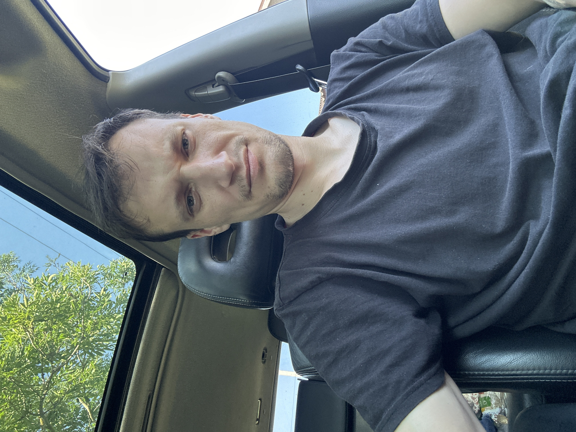 The image is a close-up, over-the-shoulder selfie of a man inside a car. He is wearing a black t-shirt and looking directly at the camera with a neutral expression. The interior of the car is visible, including the black leather seats, the headrest, and part of the dashboard. The car has a sunroof, through which green tree branches and a blue sky are visible. The image appears to have been taken during the daytime. There is no indication of the specific location within the city of Makiv, Ukraine, or any contextual details about the man's activities or the purpose of the photograph.