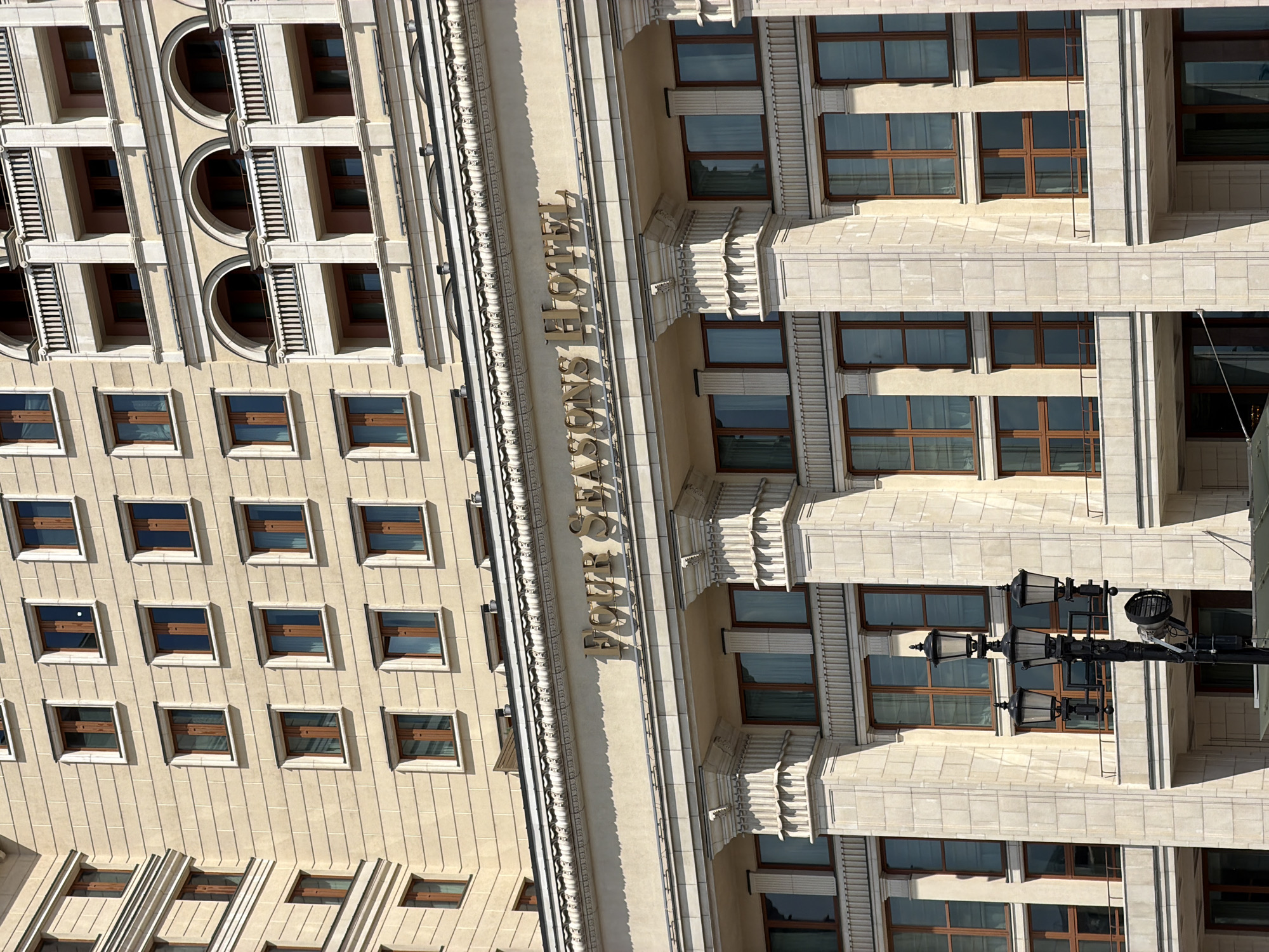 The image shows the facade of the Four Seasons Hotel in Moscow, Russia. The architecture features multiple stories with rows of windows and decorative elements. The hotel's name is displayed on the building's exterior. A lamppost is visible in the foreground.