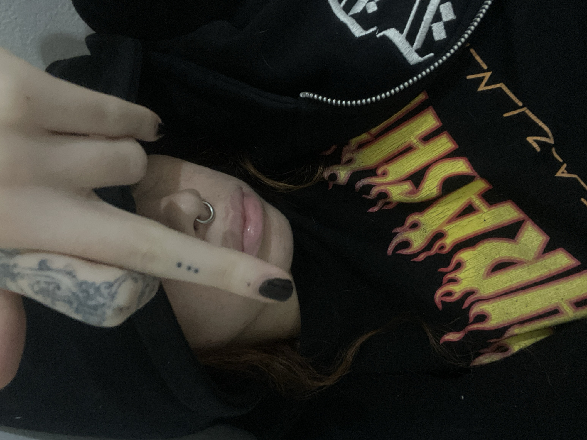 A person is lying down, wearing a black hoodie with a yellow and orange flame design on the front. They have dark nail polish and tattoos on their hand and arm. The person has a nose ring and reddish-brown hair. No specific location in Buenos Aires, Argentina, is discernible from the image.