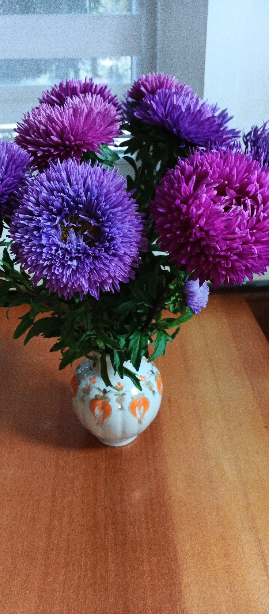 A bouquet of purple and pink asters is arranged in a white ceramic vase adorned with orange floral patterns. The vase sits on a wooden table, with a window and blinds visible in the background. The scene is indoors, likely in a home or office setting. There is no information in the image to suggest a location in Smila, Ukraine.