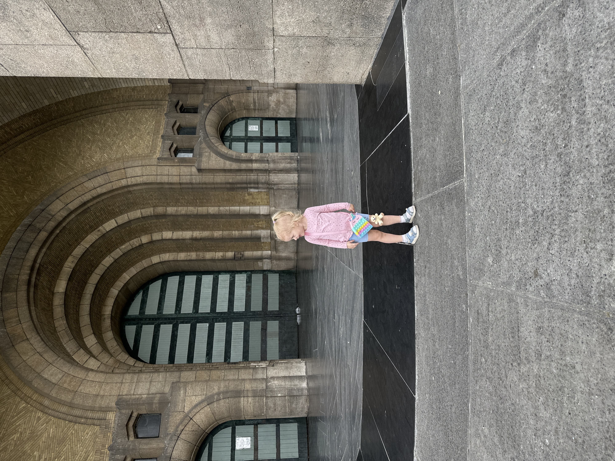 A young child with blonde hair, wearing a pink long-sleeved shirt, blue shorts, and sandals, stands in front of a large, ornate stone building with arched entrances and dark green doors. The child is holding a colorful toy in their right hand and a small plush bear is attached to their shorts. The ground in front of the building is paved with large stone slabs. The building is likely part of the National Basilica of the Sacred Heart in Koekelberg, Belgium, known for its distinctive Art Deco architecture. The scene appears to be an outdoor public space, possibly a plaza or esplanade, with the imposing facade of the basilica dominating the background. The lighting suggests it is daytime.