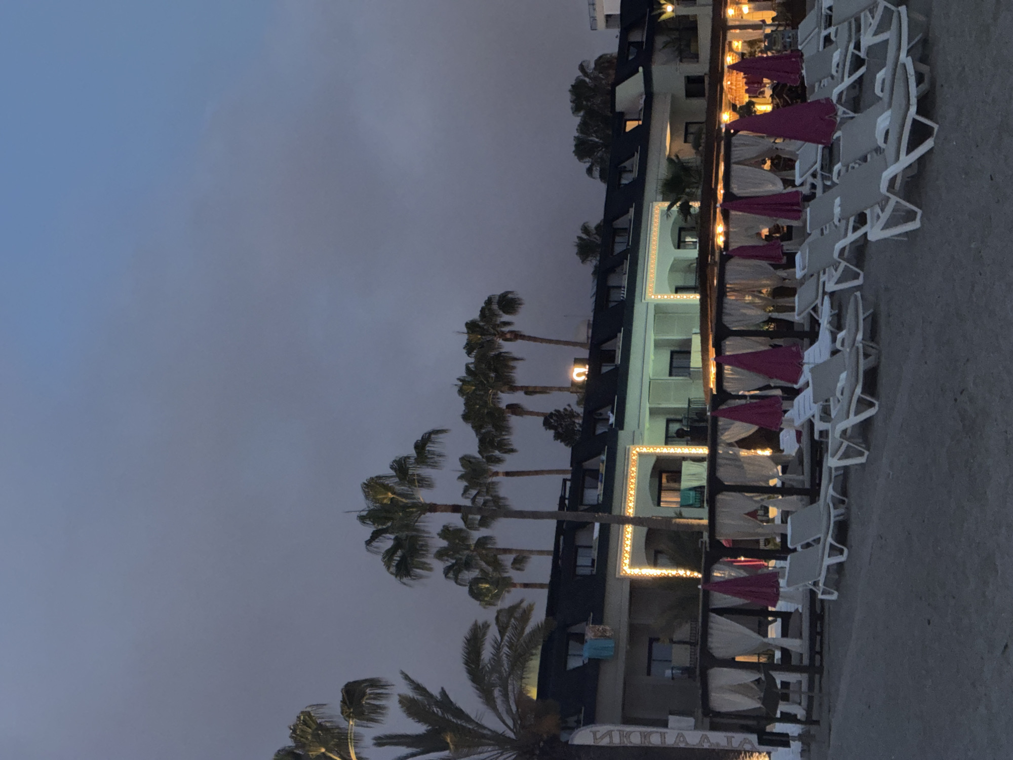 The image depicts a beach resort in Alanya, Turkey, during twilight. Several palm trees stand in front of a multi-story building adorned with decorative lights. In the foreground, beach chairs and umbrellas are arranged on the sand, with draped cabanas visible behind them. The sky is overcast, casting a dim light over the scene. No people are clearly visible in the image.